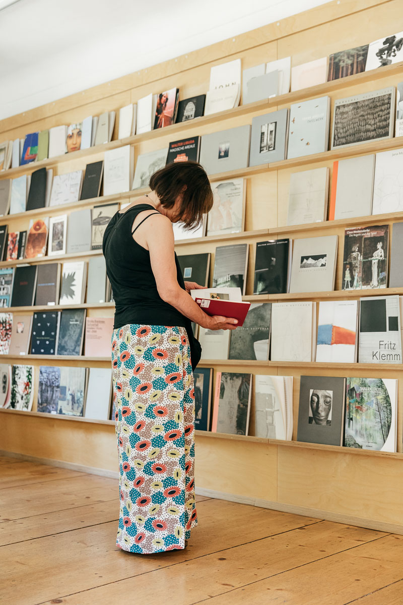 KI generiert: Eine Person steht vor einem Regal mit vielen Büchern und betrachtet eines davon. Der Raum wirkt wie eine Bibliothek oder ein Buchladen.