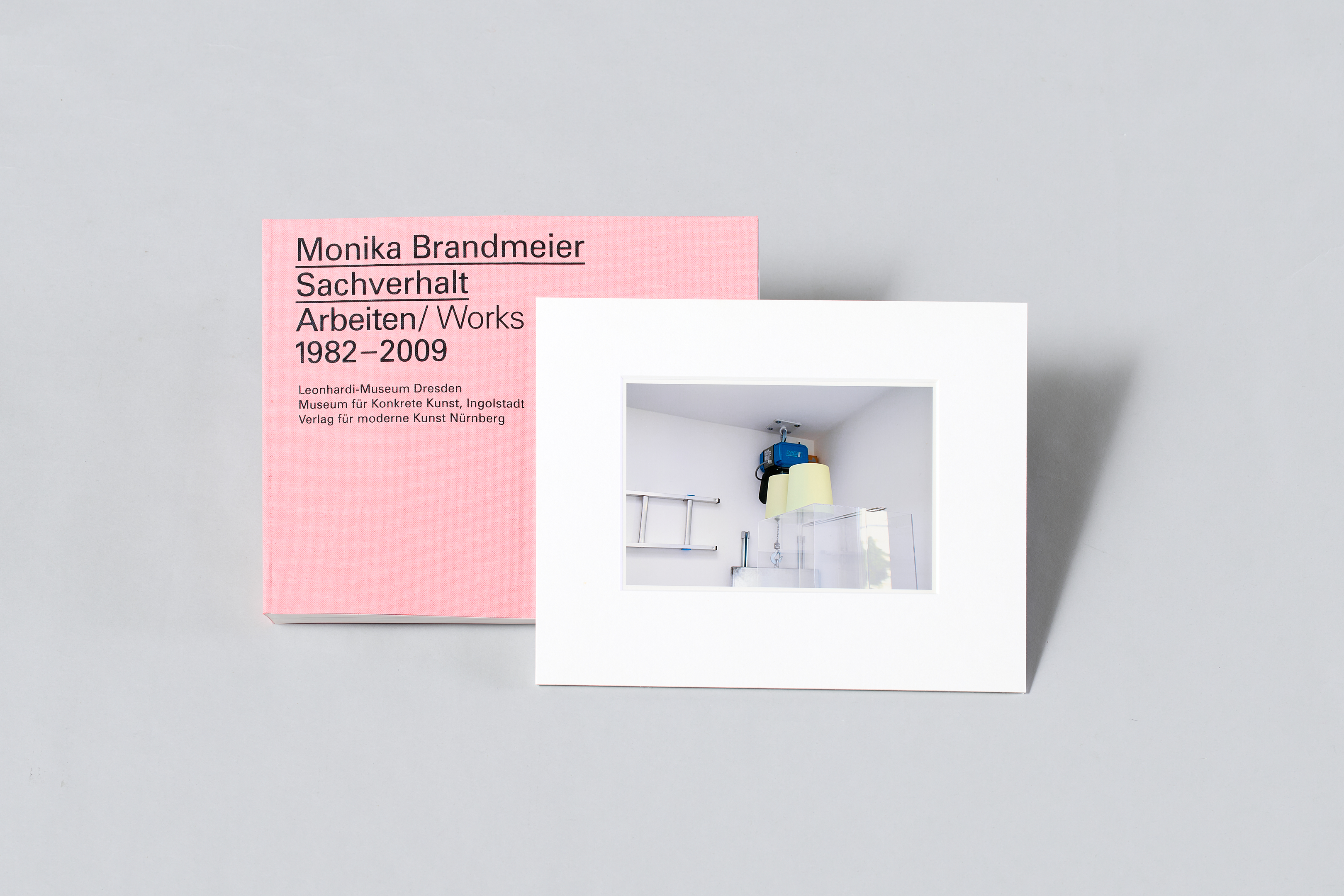 KI generiert: Auf dem Bild ist ein Buch mit dem Titel "Monika Brandmeier Sachverhalt Arbeiten/Works 1982–2009" in einer rosa Abdeckung zu sehen, daneben liegt ein weißes Passepartout mit einem Foto. Das Foto zeigt ein minimalistisches Kunstwerk oder eine Installation in einem Raum.