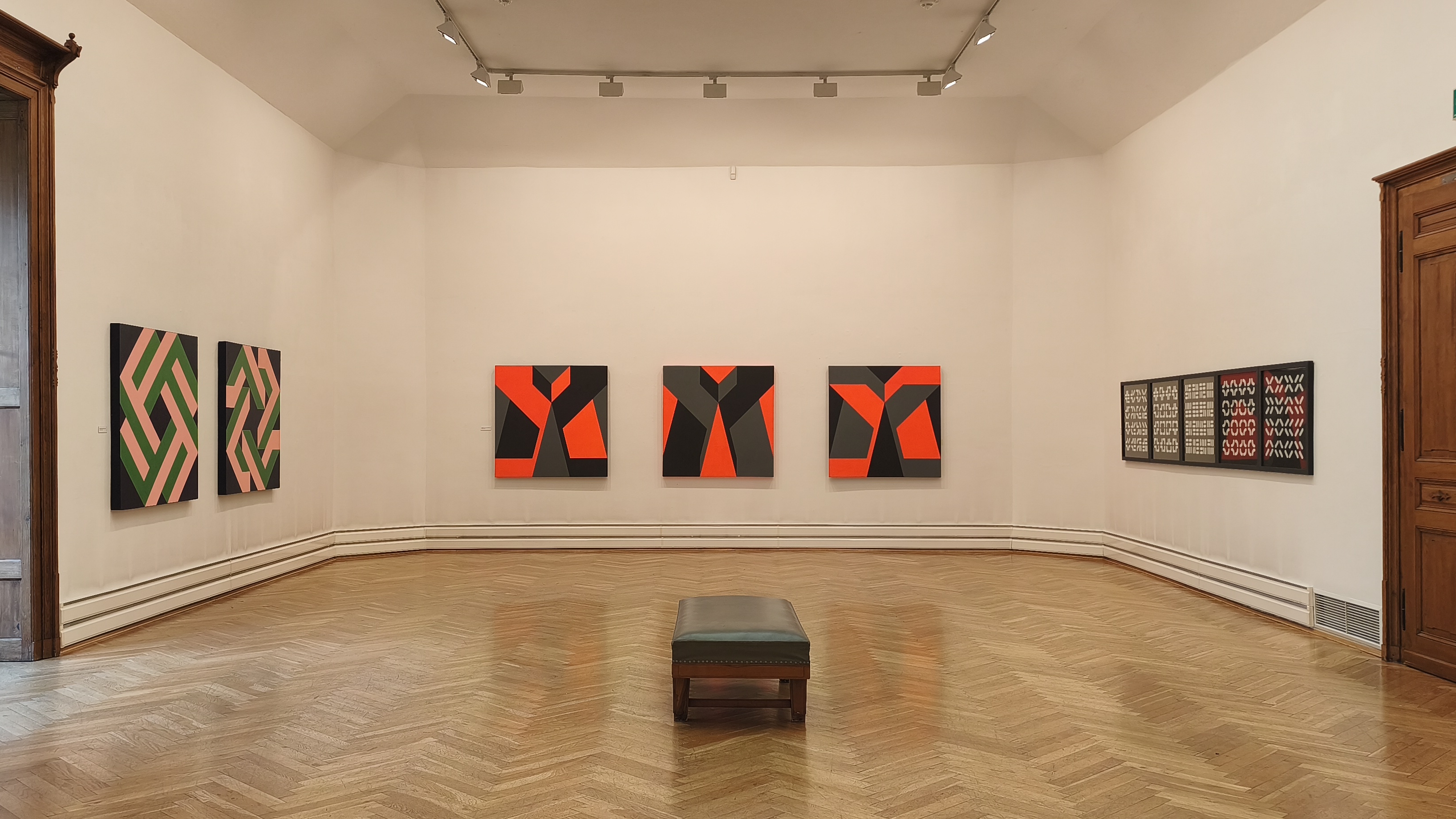 KI generiert: Das Bild zeigt einen Galerie- oder Museumsraum mit geometrischen Kunstwerken an den Wänden, hauptsächlich in Schwarz, Rot und Grün gehalten. In der Mitte des Raumes steht eine schlichte Sitzbank auf einem Holzfußboden.