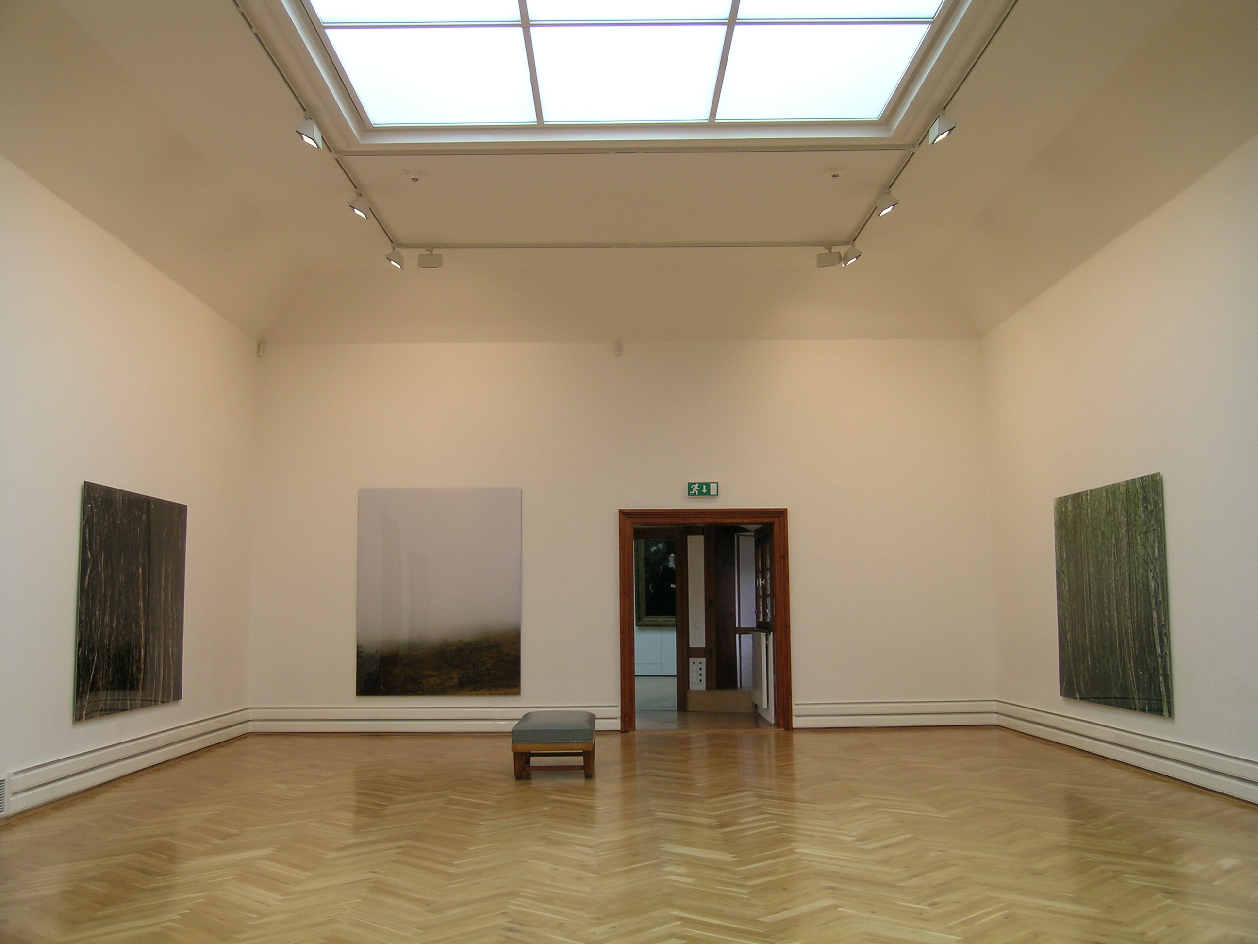 KI generiert: Das Bild zeigt einen leeren Kunstgalerieraum mit drei großformatigen Bildern an den Wänden und einer Bank in der Mitte. Durch eine offene Tür ist ein Teil eines weiteren Raumes sichtbar.