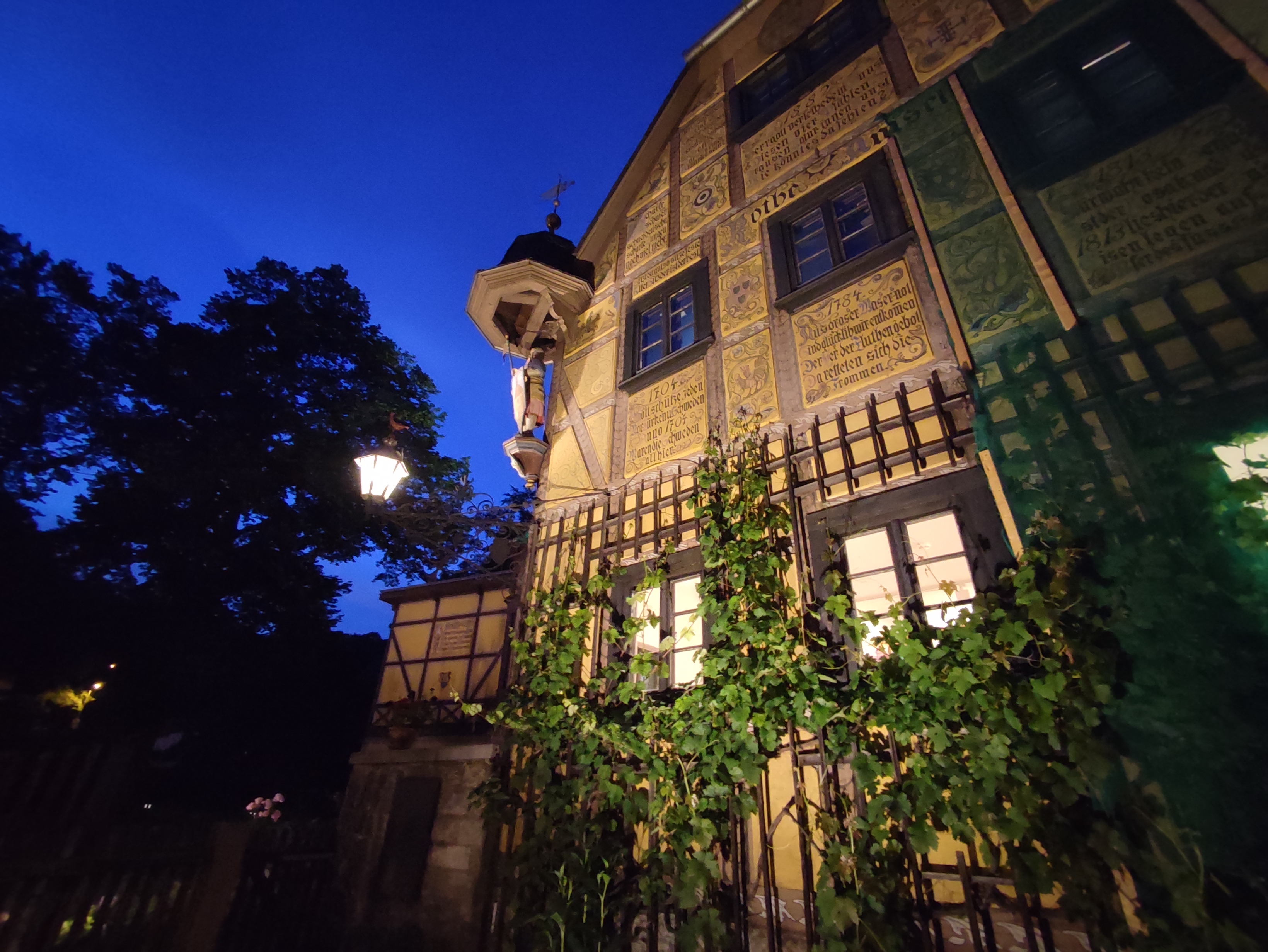 KI generiert: Das Bild zeigt ein historisches Fachwerkhaus bei Nacht, das mit Beschriftungen und Schnitzereien verziert ist. Im Vordergrund wächst Efeu an der Fassade, während eine Straßenlaterne und der dunkelblaue Himmel eine stimmungsvolle Atmosphäre schaffen.