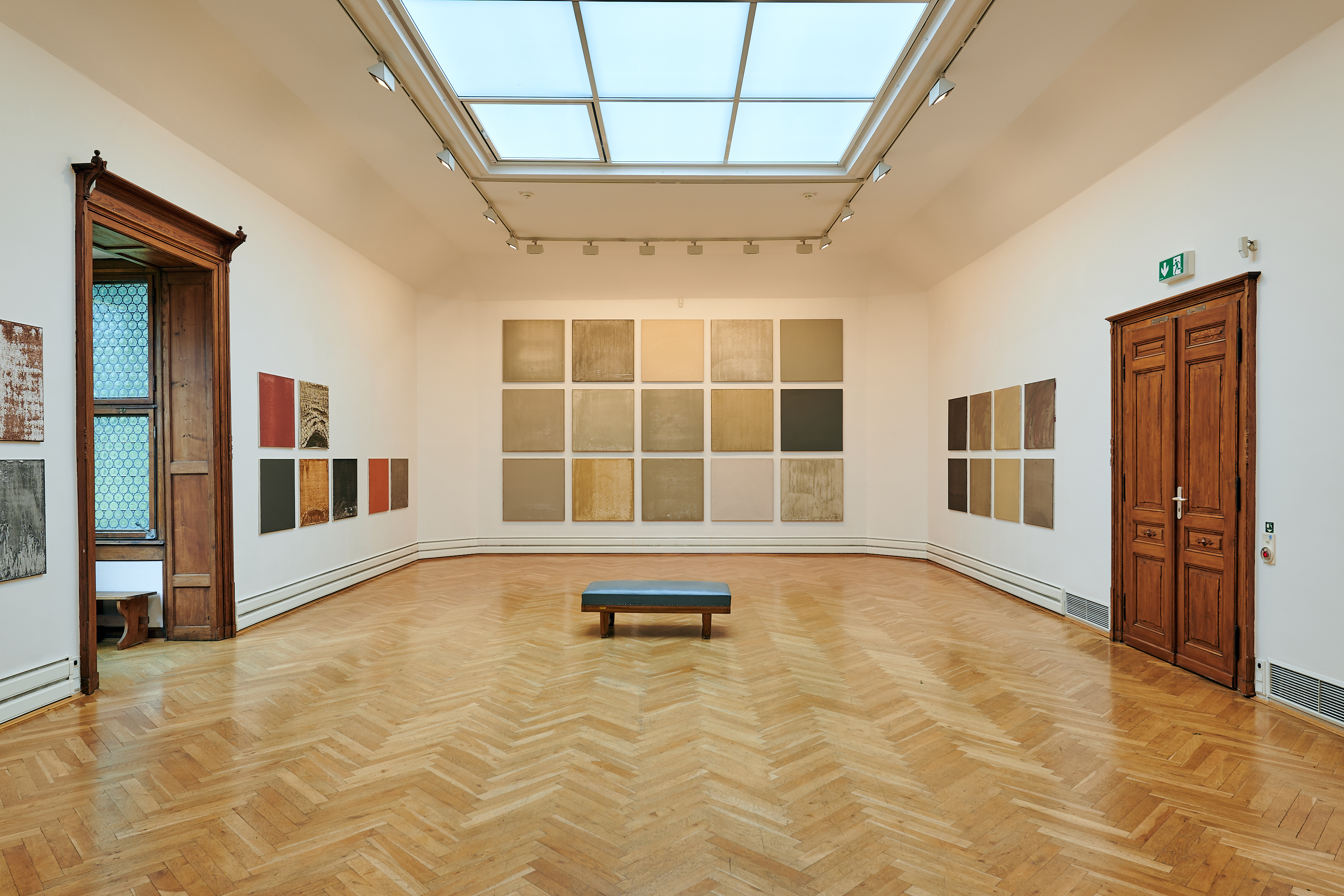 KI generiert: Das Bild zeigt eine Galerie mit minimalistischen, abstrakten Kunstwerken an den Wänden und Parkettboden. Im Zentrum des Raums steht eine Bank, über der sich ein großes Oberlicht befindet.