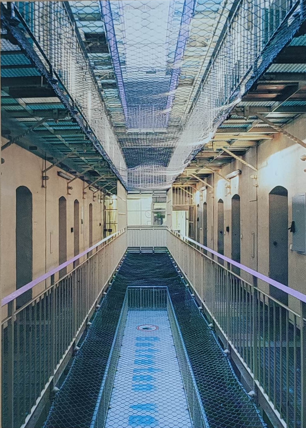 KI generiert: Das Bild zeigt den Innenraum eines Gefängnisses mit Zellen auf beiden Seiten und Sicherheitsnetzen an Decken und Böden. Die Struktur ist hell erleuchtet und symmetrisch aufgebaut.