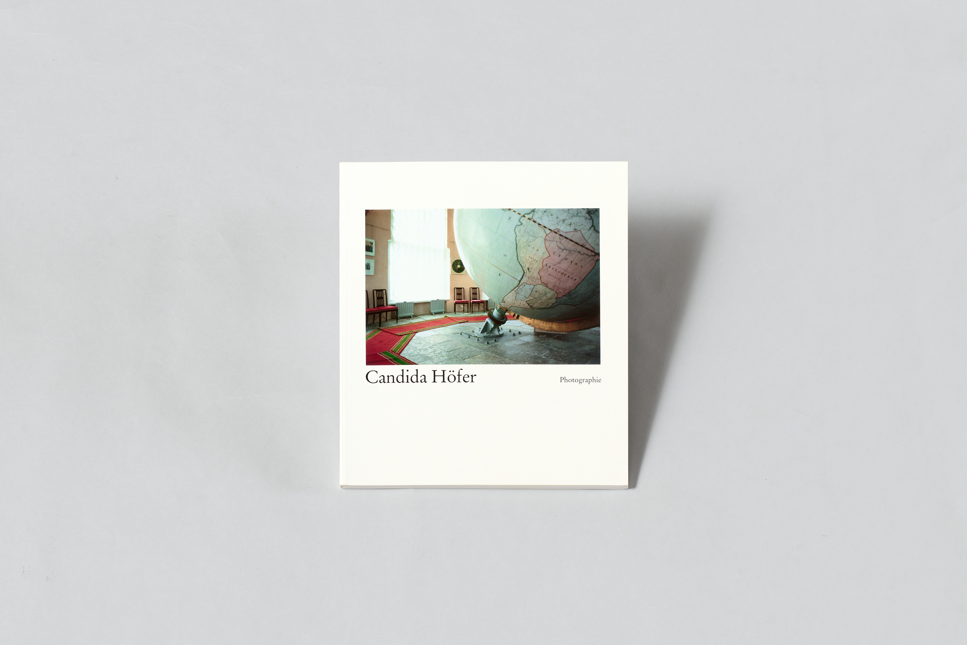 AI generated: The image shows the cover of a book by Candida Höfer with a photo of a person next to a large globe in a room. The artist's name can be seen on the cover.