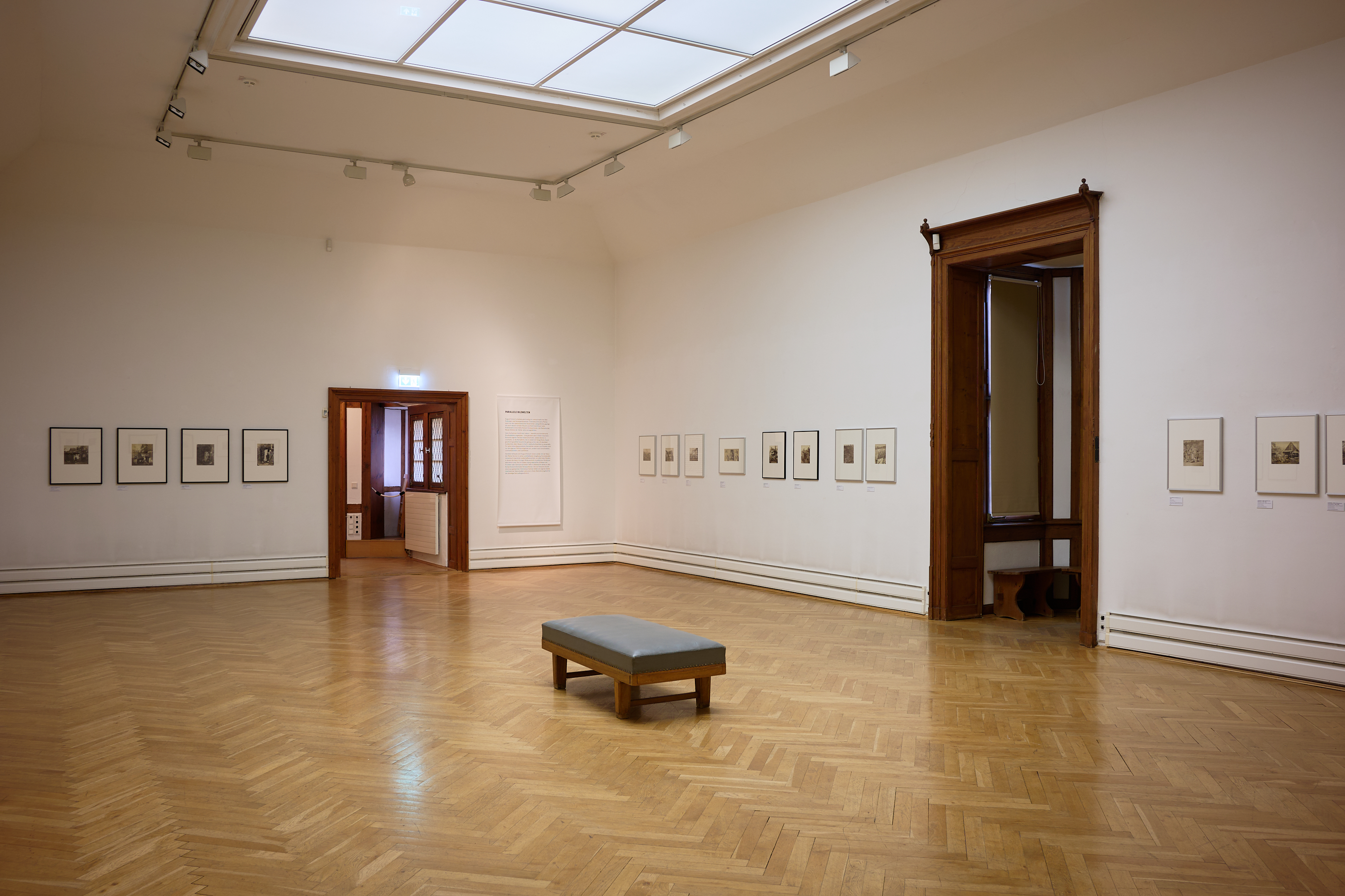 AI generated: The image shows an empty museum room with a wooden floor, in which numerous framed works of art hang on the walls. There is a single grey bench in the middle of the room.