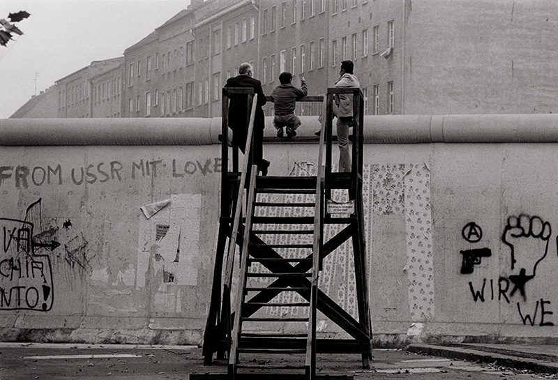 KI generiert: Das Bild zeigt drei Menschen, die auf einer Holzkonstruktion sitzen und über die Berliner Mauer blicken. An der Mauer sind Graffitis mit politischen Botschaften zu sehen.