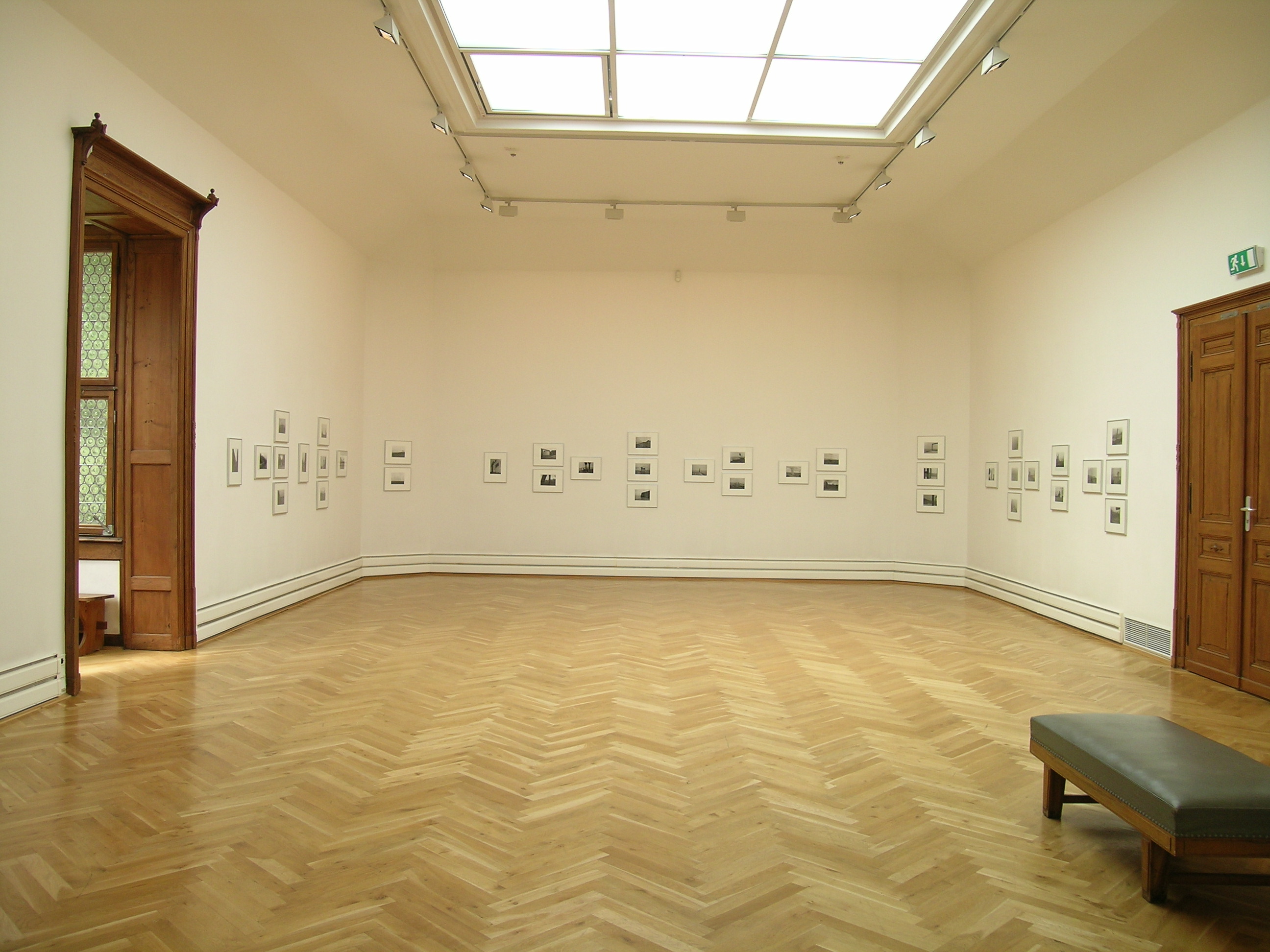 KI generiert: Das Bild zeigt einen hellen und minimalistischen Galerieraum mit Holzboden, weißen Wänden und kleinen gerahmten Kunstwerken, die an den Wänden hängen. Zwei Türen und eine Bank sind ebenfalls im Raum zu sehen.