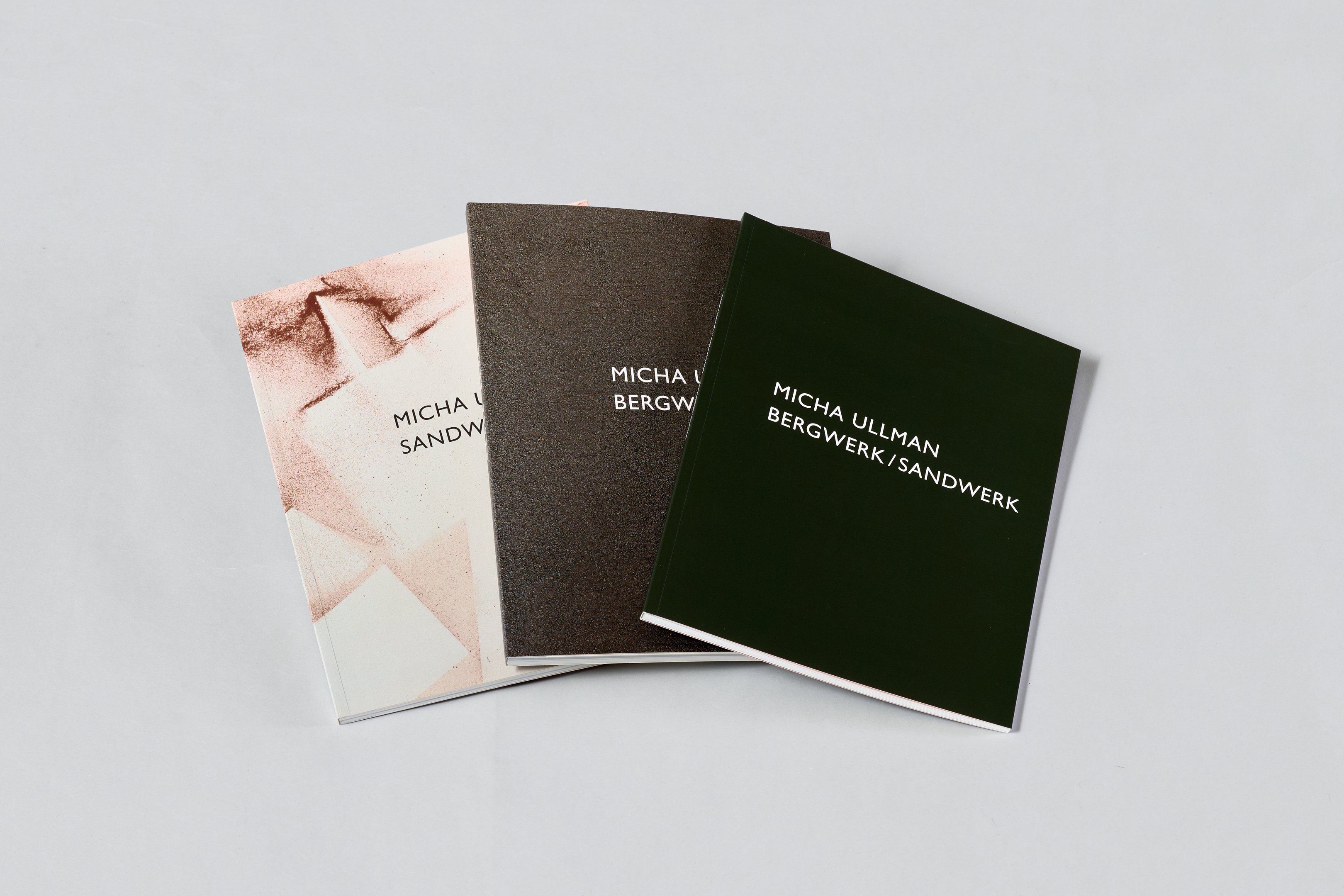 AI generated: The image shows three books with the title "Micha Ullman: Bergwerk/Sandwerk" lying next to each other on a flat surface. Each book has a unique cover design.