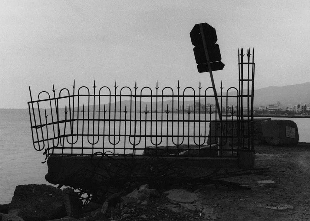 KI generiert: Das Bild zeigt ein beschädigtes Metallzaun am Ufer, daneben ein verbogenes Verkehrsschild. Im Hintergrund ist das Wasser und eine Stadtlandschaft zu sehen.