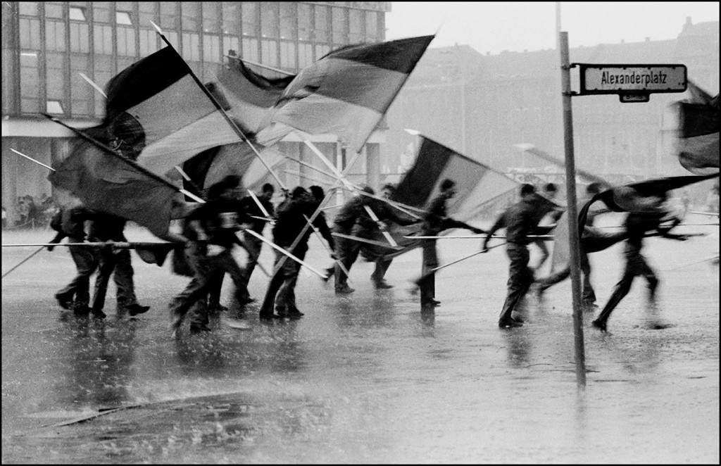 KI generiert: Das Bild zeigt eine Gruppe von Menschen, die bei Regen mit großen Fahnen über einen Platz laufen, wobei im Vordergrund ein Straßenschild mit der Aufschrift "Alexanderplatz" zu sehen ist. Hauptinhalt ist die Bewegung der Personen und der nasse, regnerische Zustand.