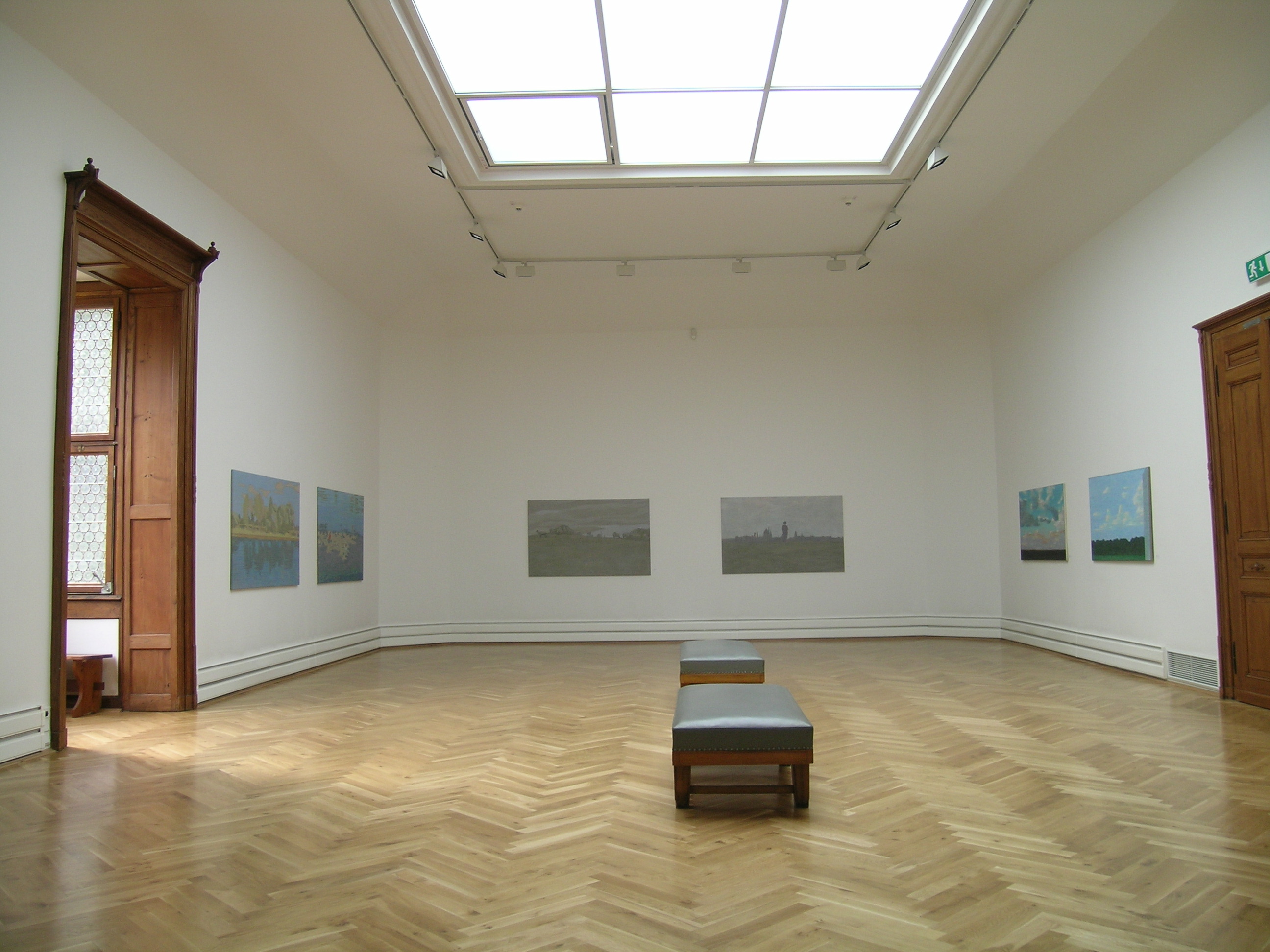 KI generiert: Das Bild zeigt einen hellen und geräumigen Ausstellungsraum mit Gemälden an den Wänden und Bänken in der Mitte des Raumes. Ein großes Glasdach lässt viel Licht herein, und die Holztüren und der Parkettboden verleihen dem Raum eine klassische Atmosphäre.