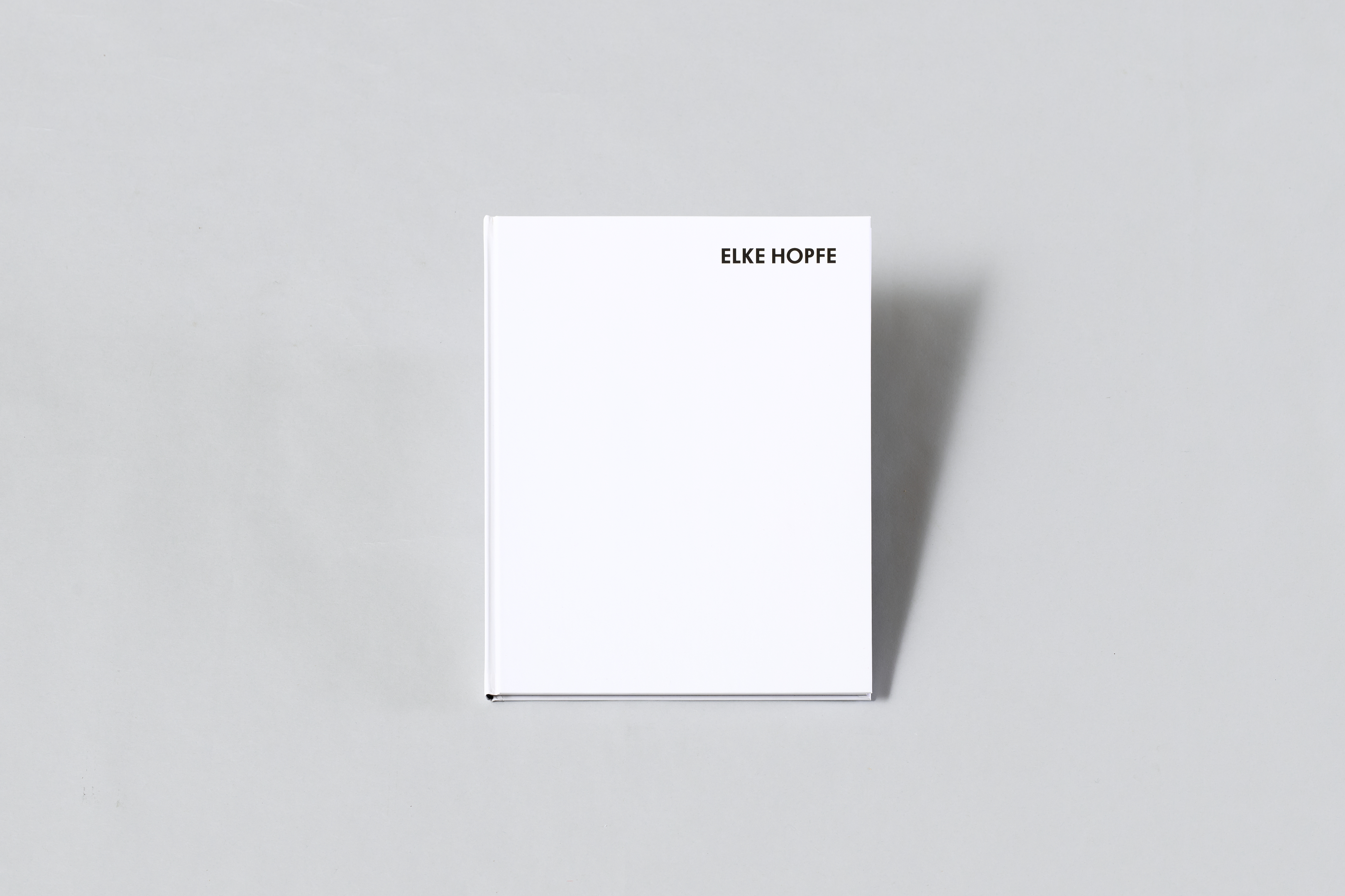 AI generated: The image shows a plain, white book with the name "Elke Hopfe" on the cover. It lies on a neutral, grey surface.