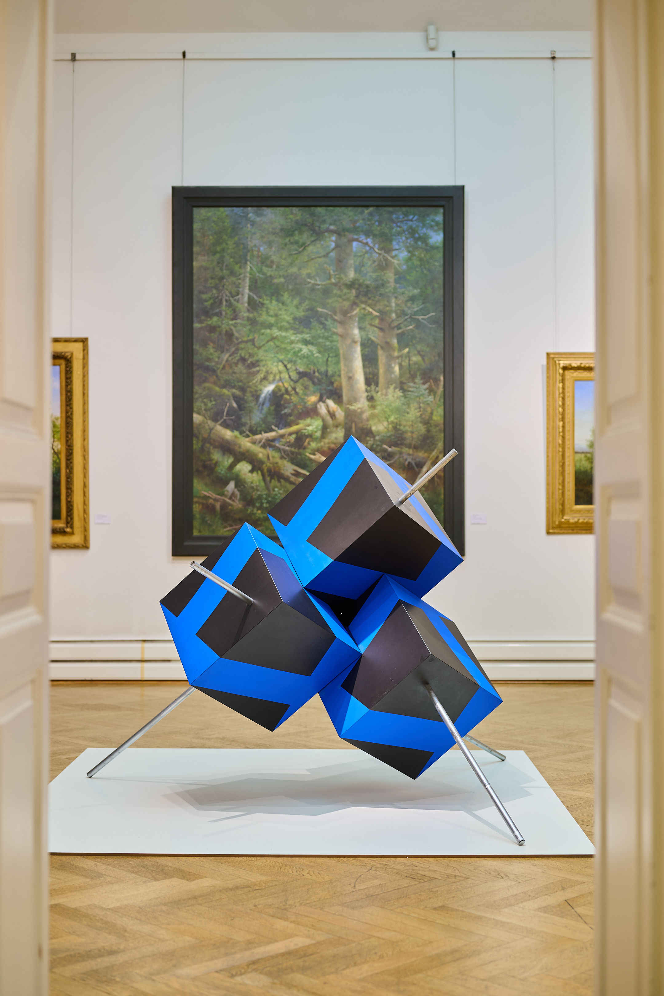 KI generiert: Das Bild zeigt eine moderne Skulptur aus blauen und schwarzen geometrischen Formen, die auf Metallstangen balanciert. Im Hintergrund hängen klassische Gemälde an einer Galerie-Wand.