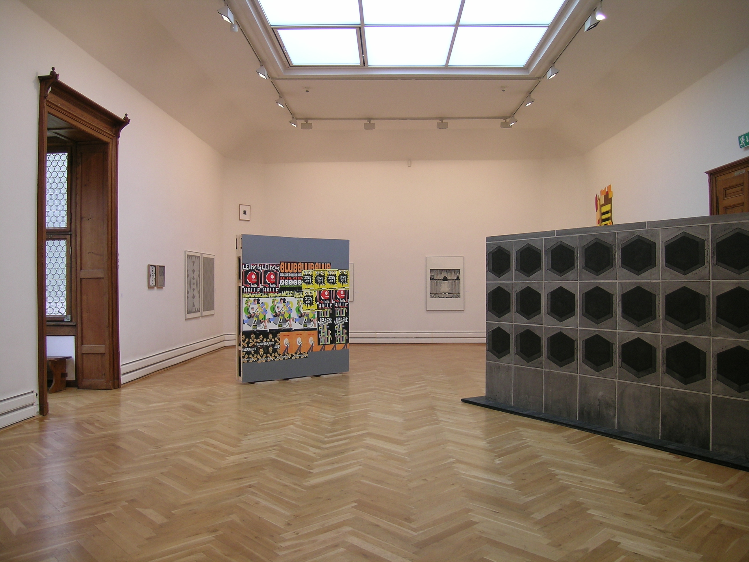 KI generiert: Das Bild zeigt einen modernen Ausstellungsraum mit Holzboden, einem großen Oberlicht und mehreren Kunstwerken, darunter ein gemaltes Bild und eine strukturierte Skulptur. Der Raum hat helle Wände und ist minimalistisch gestaltet.