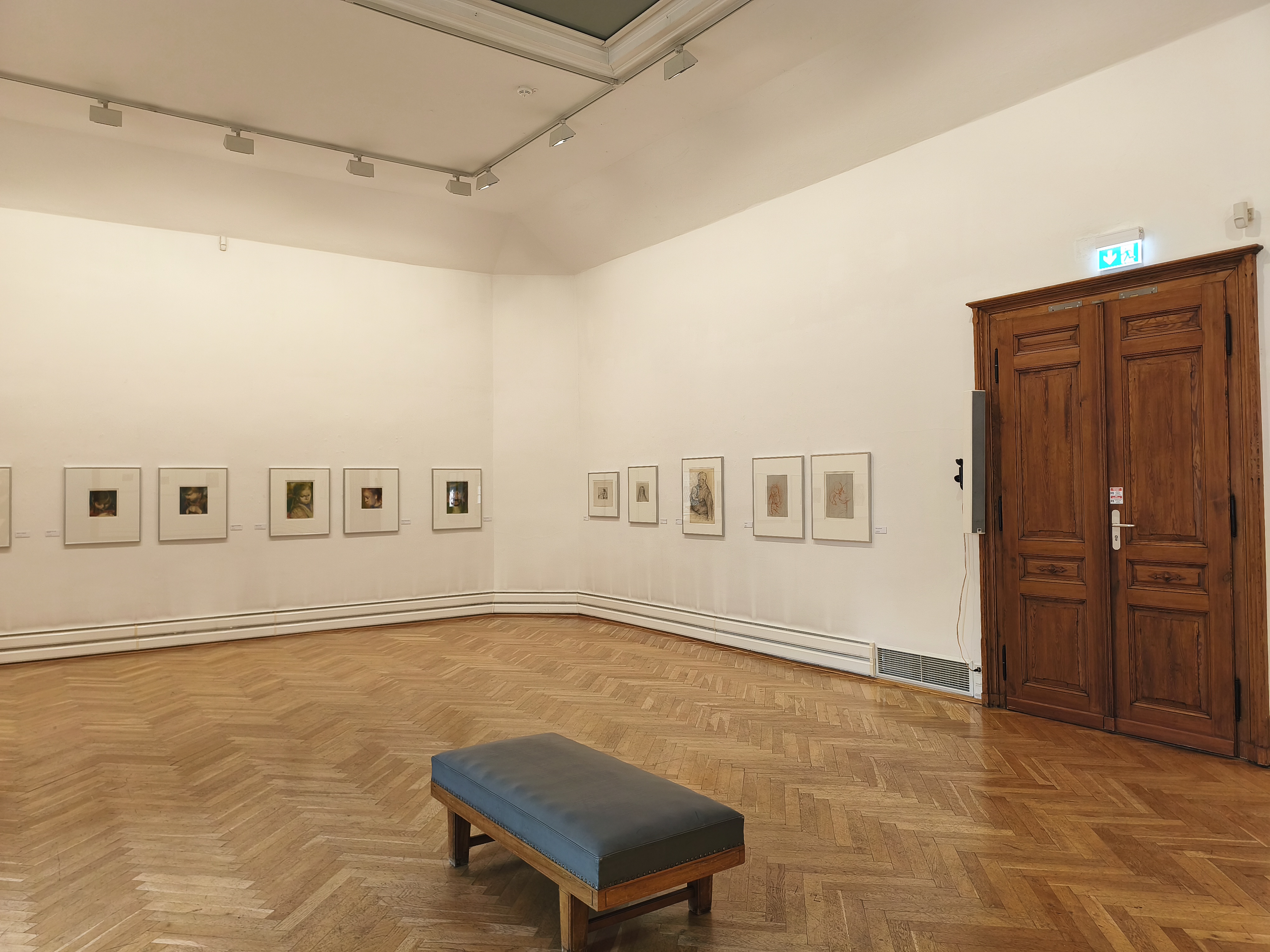 KI generiert: Das Bild zeigt einen Museumsraum mit hölzernem Parkettboden. An den Wänden hängen mehrere gerahmte Kunstwerke, und in der Mitte steht eine gepolsterte Sitzbank.