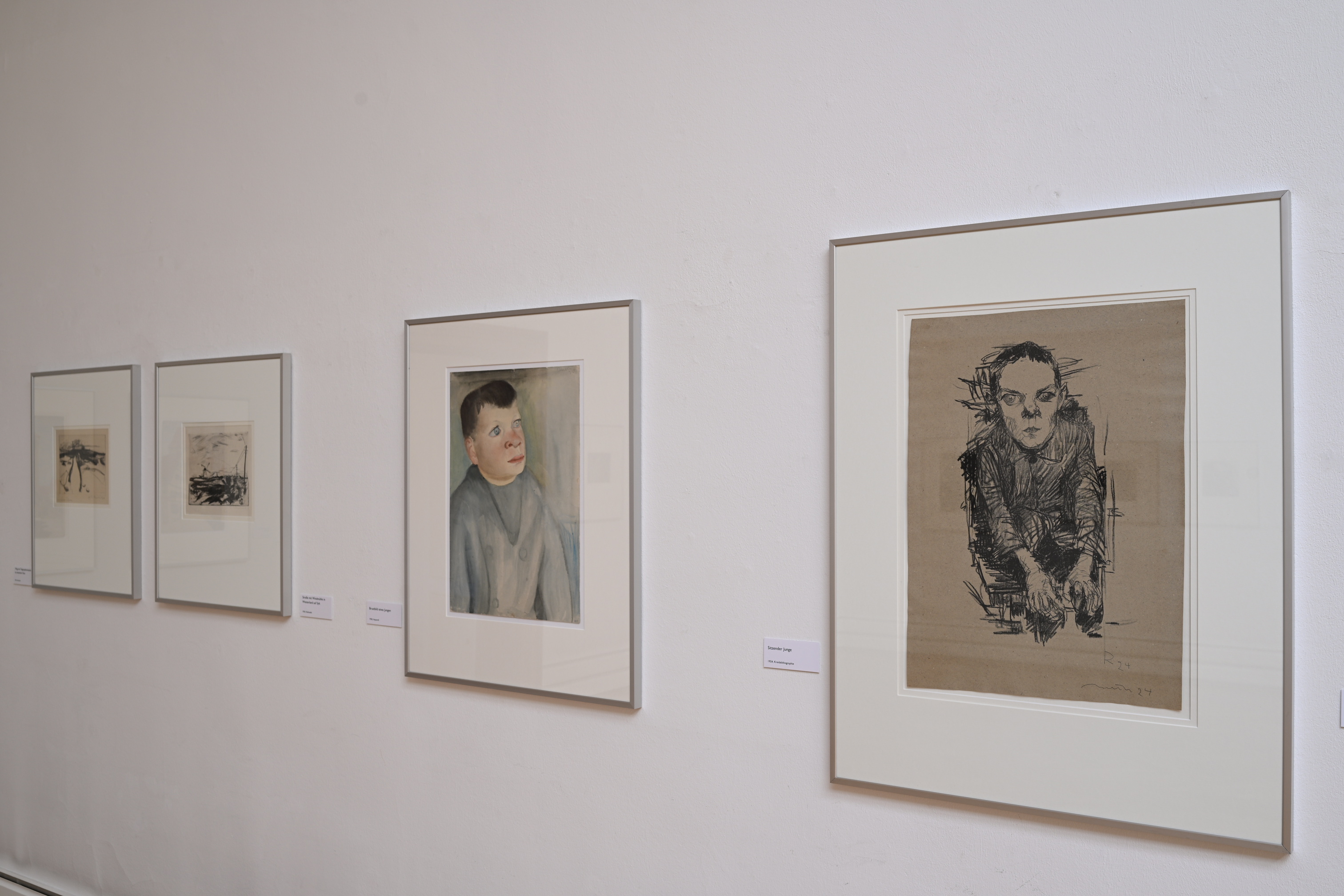 KI generiert: Das Bild zeigt eine Ausstellung von Kunstwerken, die in weißen Rahmen an einer Wand hängen. Die Werke beinhalten Zeichnungen und Malereien von Personen in unterschiedlichen Stilen.