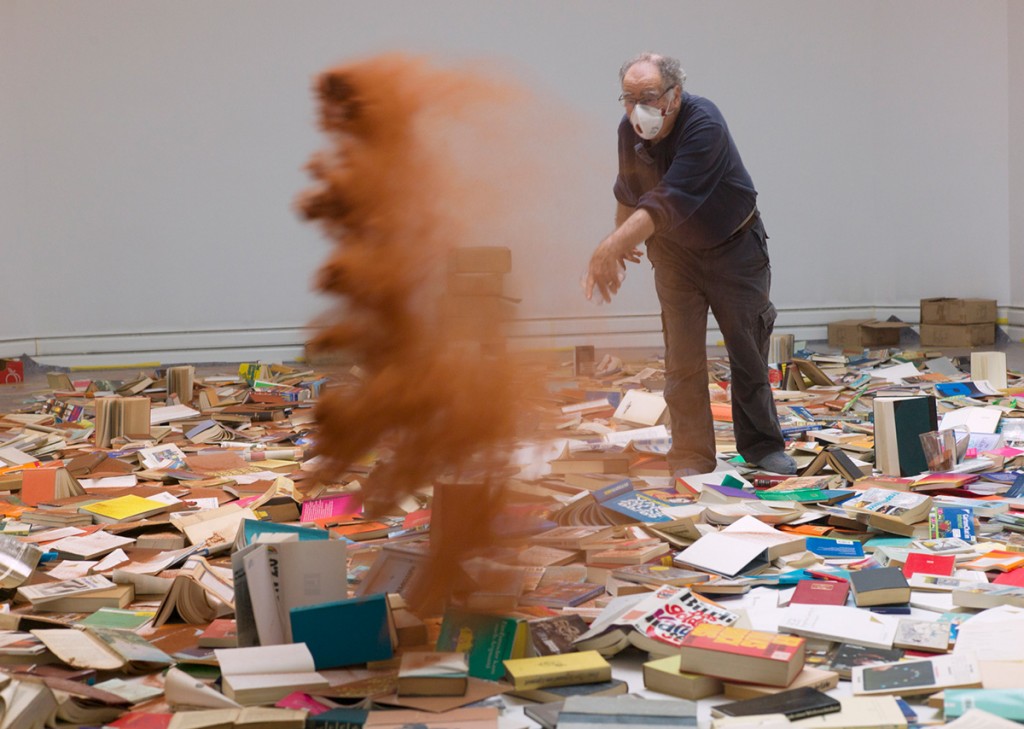 KI generiert: Ein Mann mit einer Schutzmaske wirft, in einem Raum voller verstreuter Bücher, eine Wolke aus rötlichem Staub in die Luft.