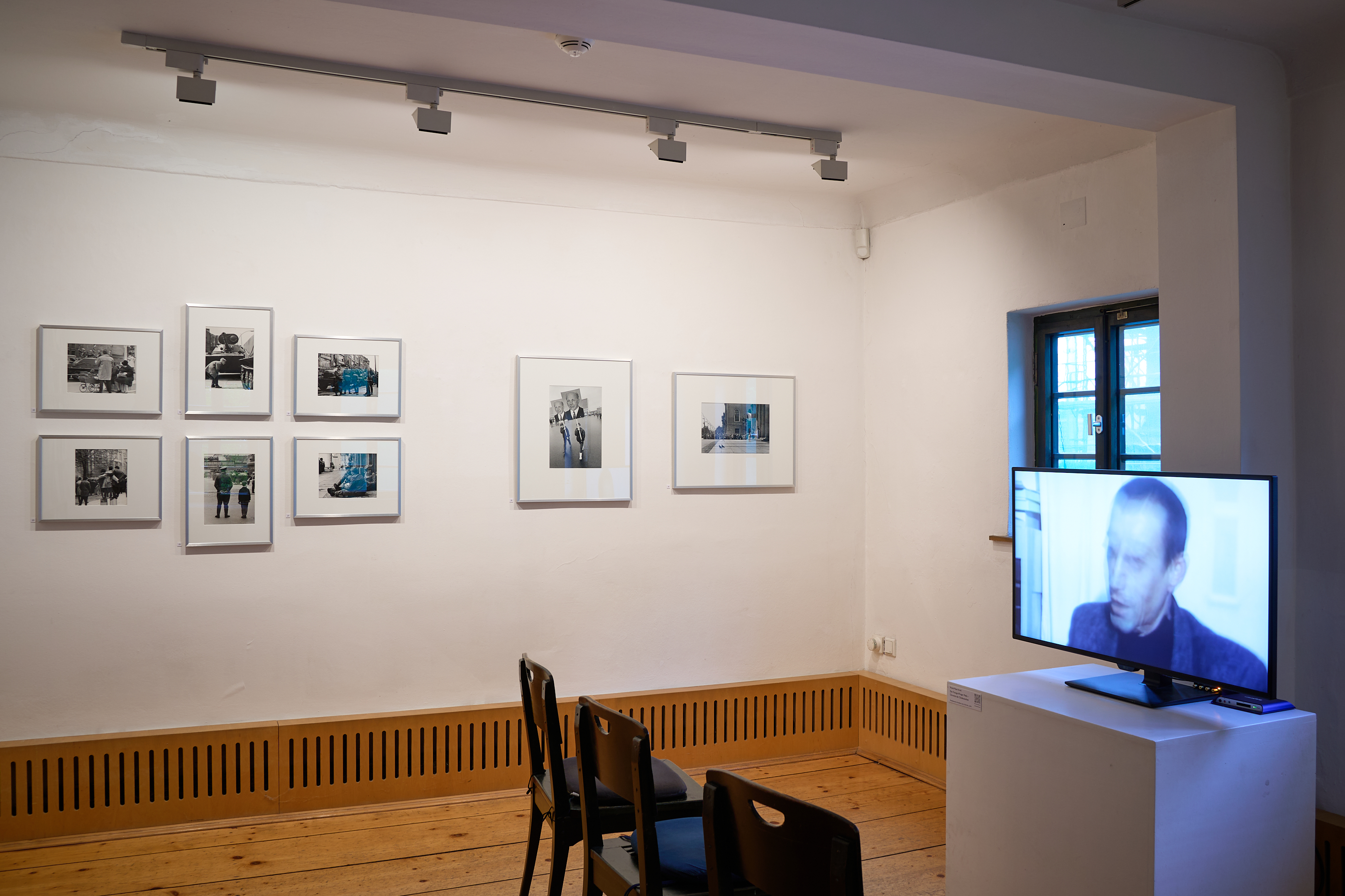 KI generiert: Das Bild zeigt einen Ausstellungsraum mit an der Wand hängenden Fotografien und einem Monitor mit Videoprojektion. Zwei Stühle sind vor dem Monitor platziert, die Exponate bieten eine moderne Präsentation.