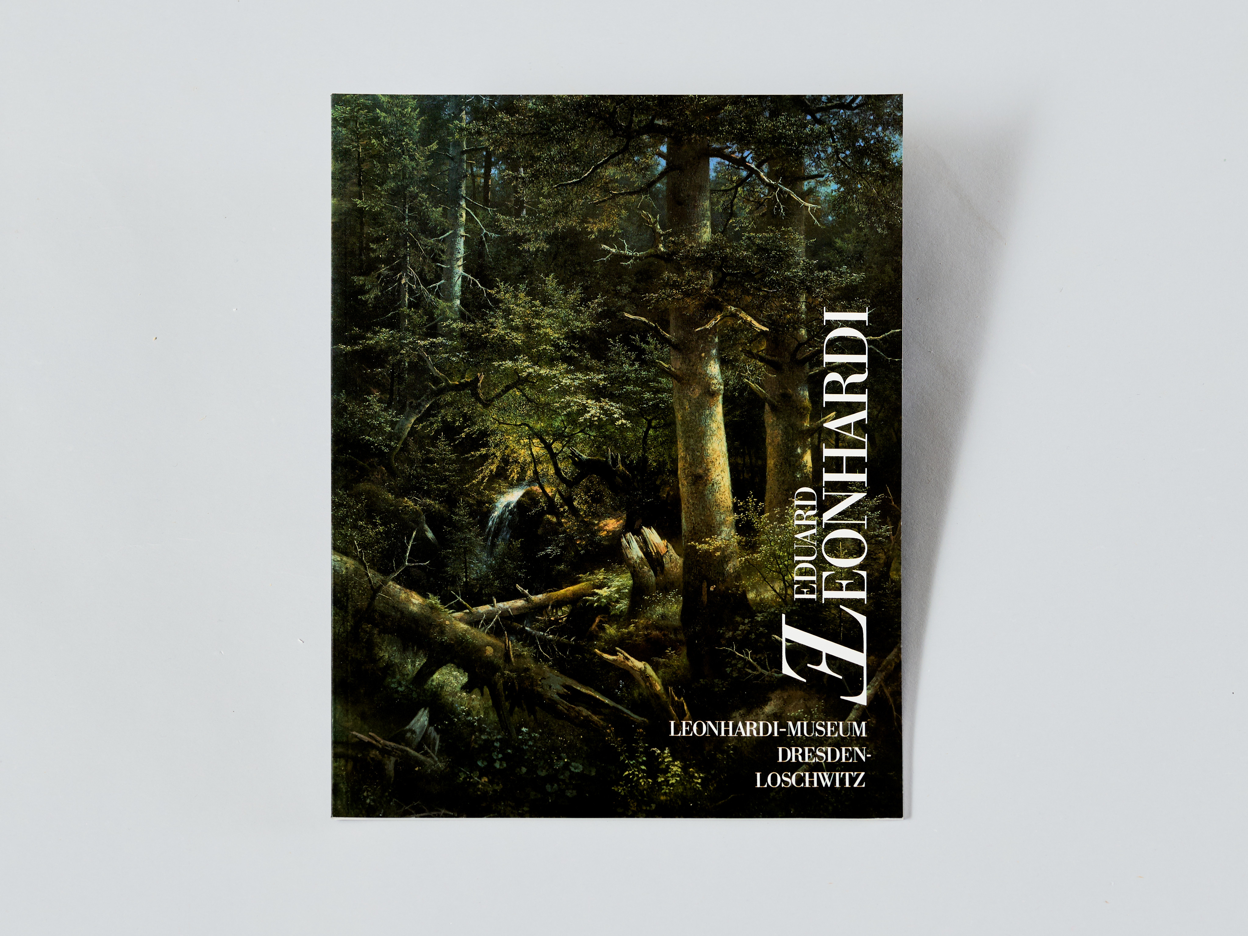 KI generiert: Das Bild zeigt ein kleines Buch oder eine Broschüre mit einem dunkelgrünen, waldähnlichen Cover-Design, das auf einem neutralen Hintergrund liegt. Auf dem Cover steht "LEONARDI."