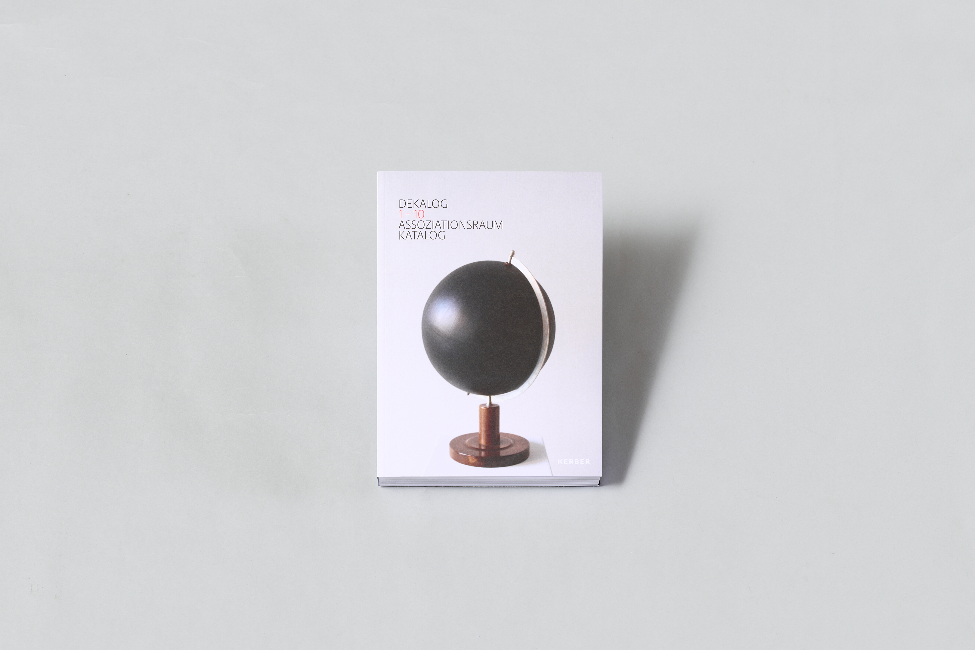 AI generated: The image shows a book with the title "DEKALOG 1-10 Assoziationsraum Katalog", on the cover of which there is an image of a black globe. The background is monochrome grey.