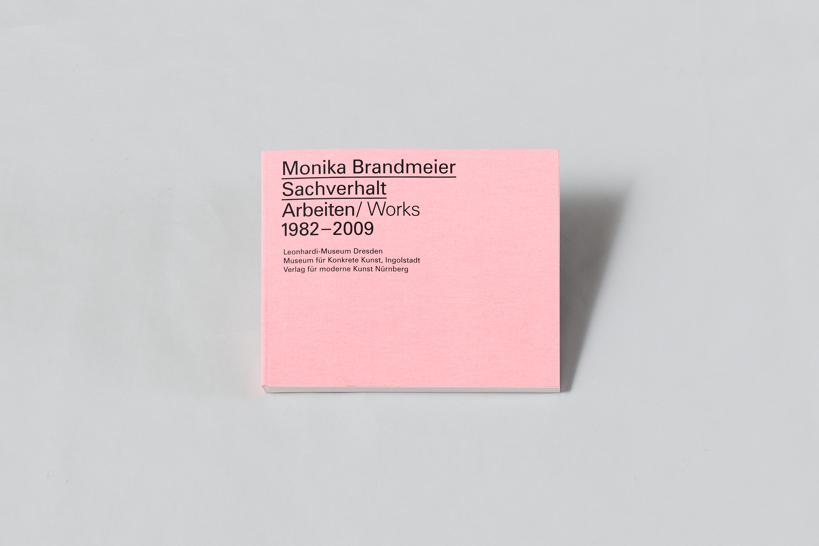 KI generiert: Das Bild zeigt ein Buch mit einem rosa Einband, auf dem der Titel "Monika Brandmeier: Sachverhalt Arbeiten/Works 1982–2009" zu sehen ist. Es handelt sich um eine Publikation über die Arbeiten der Künstlerin Monika Brandmeier.
