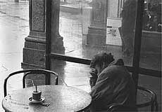 KI generiert: Das Bild zeigt eine Person mit niedergeschlagenem Kopf, die in einem Café sitzt, während es draußen regnet. Auf dem Tisch steht eine Tasse mit Untertasse.