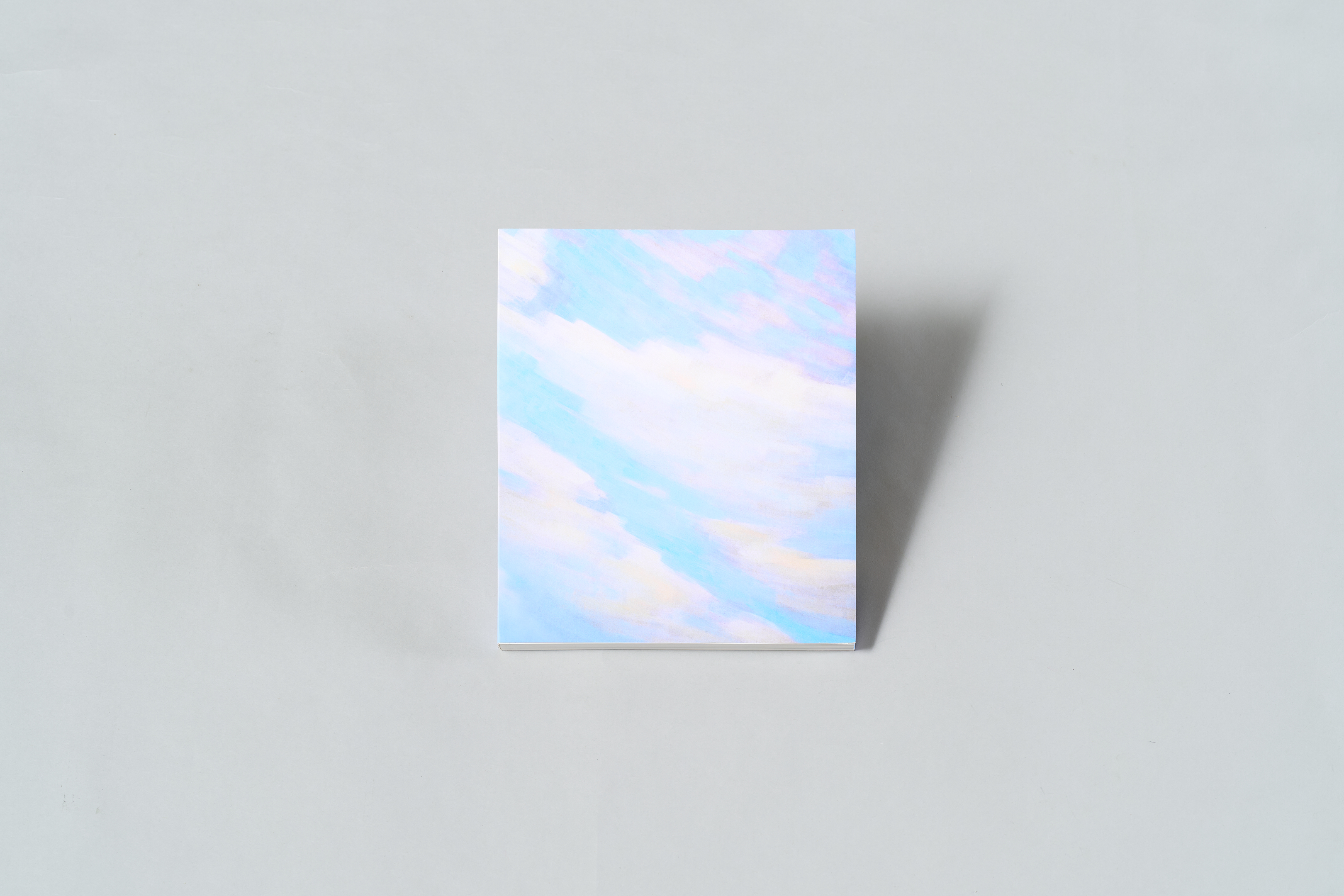 AI generated: The image shows a notebook with a pastel-coloured cover reminiscent of a sky with clouds. It lies on a neutral grey background.