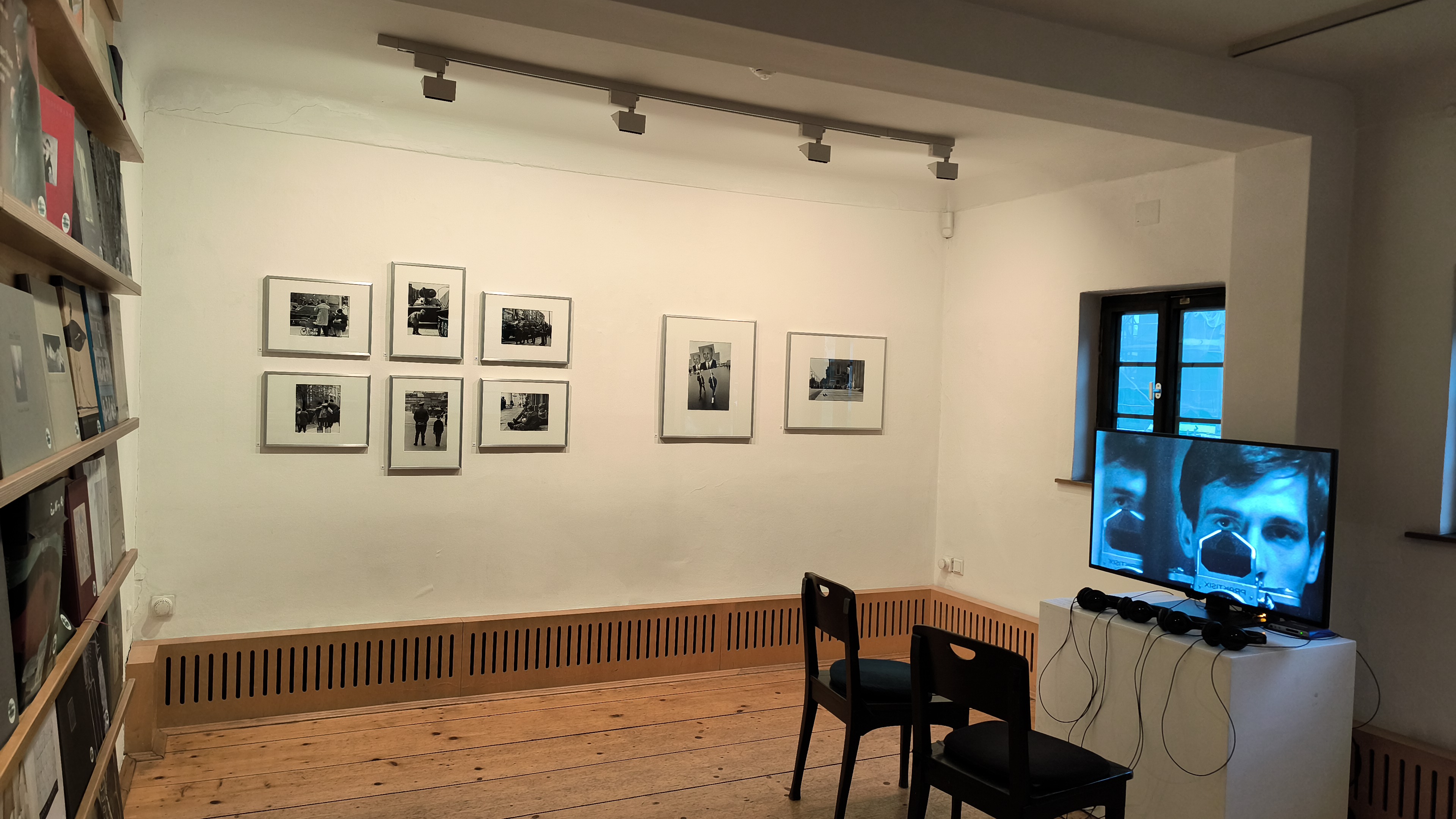 KI generiert: Das Bild zeigt eine kleine Galerie oder Ausstellung mit Schwarz-Weiß-Fotografien an der Wand und einem Monitor, der Videoaufnahmen zeigt. Im Raum sind zwei Stühle vor dem Monitor platziert.