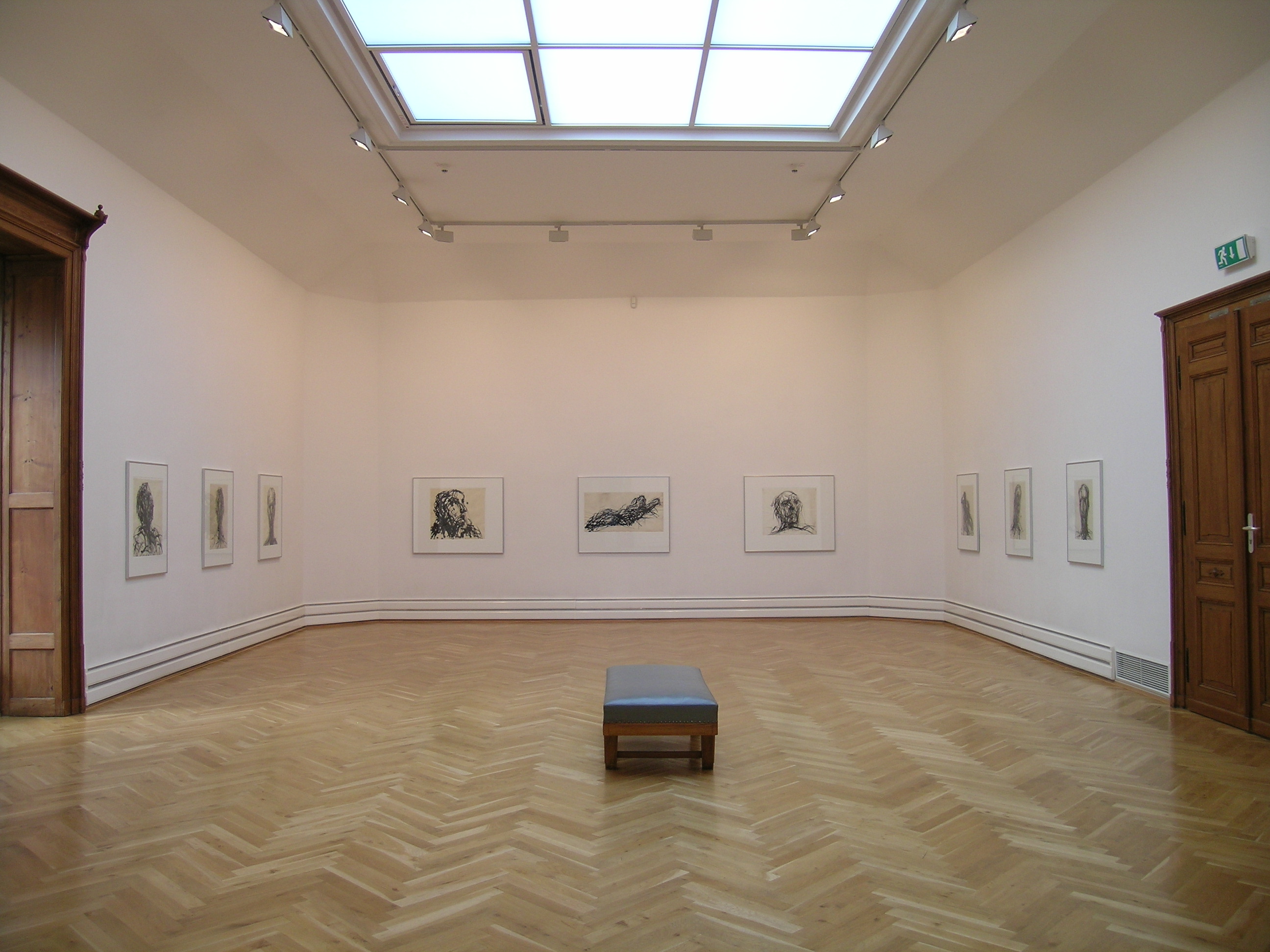 KI generiert: Das Bild zeigt einen leeren Ausstellungsraum in einer Kunstgalerie mit Skizzen an den Wänden und einer einfachen Bank in der Mitte. Die Galerie hat einen hellen Holzboden und ein großes Dachfenster, das den Raum beleuchtet.