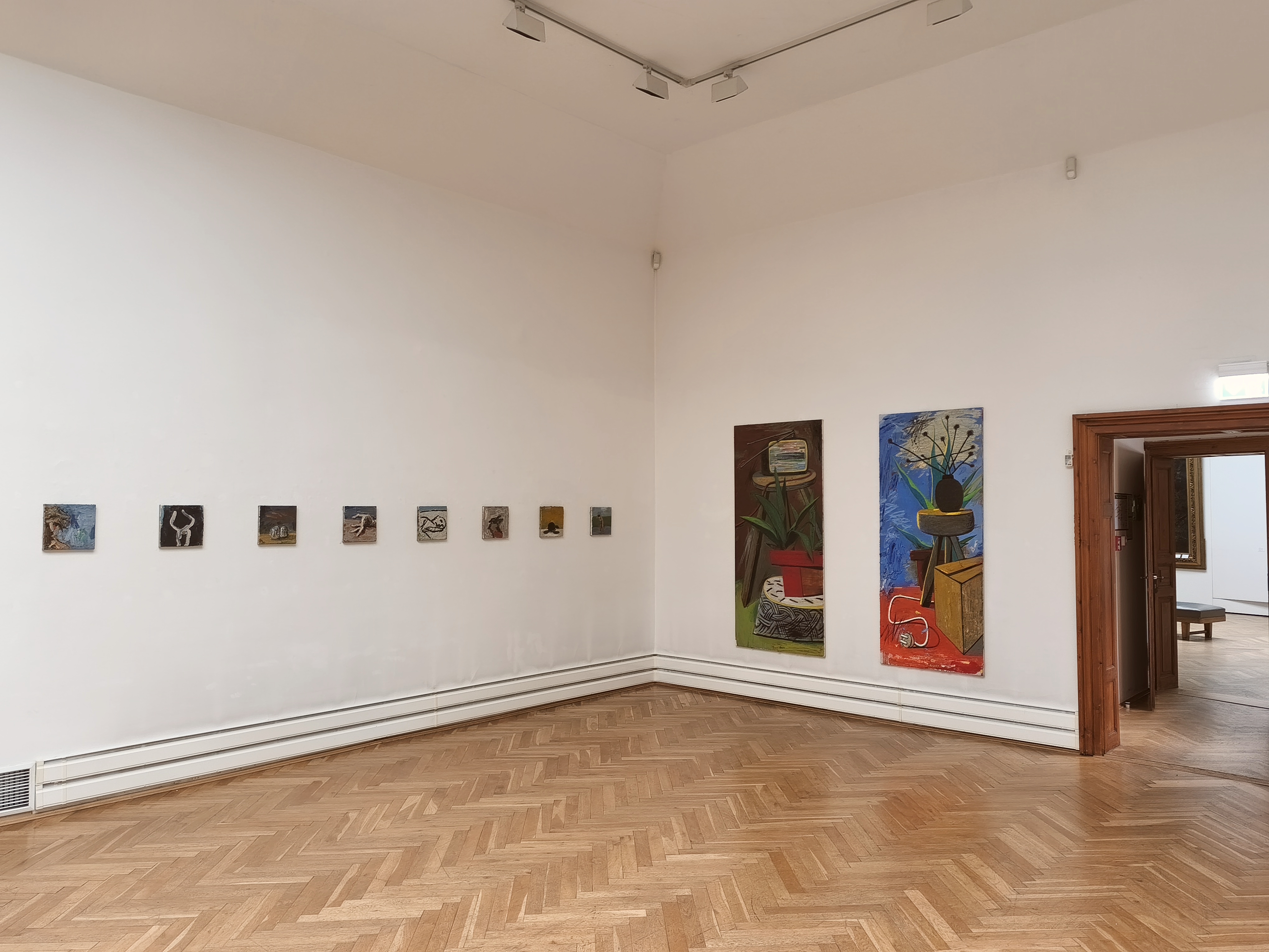 KI generiert: Das Bild zeigt eine Galerie mit mehreren kleinen und zwei großen Gemälden an weißen Wänden, die Kunstwerke in verschiedenen Stilen präsentieren. Der Raum hat einen Holzboden und es führt eine Tür in einen weiteren Bereich der Galerie.