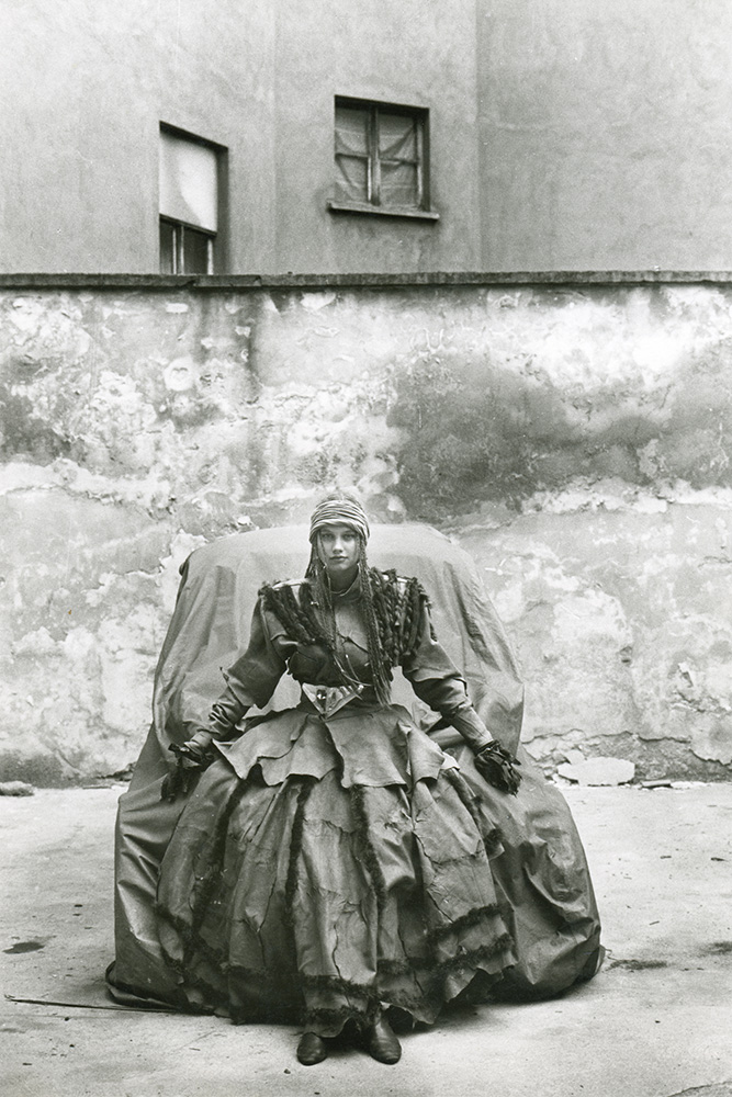 KI generiert: Das Bild zeigt eine Person in einem aufwändigen, historischen Kostüm, die vor einer verwitterten Wand auf einem Stuhl sitzt. Der Hintergrund ist schlicht und hebt die Person besonders hervor.