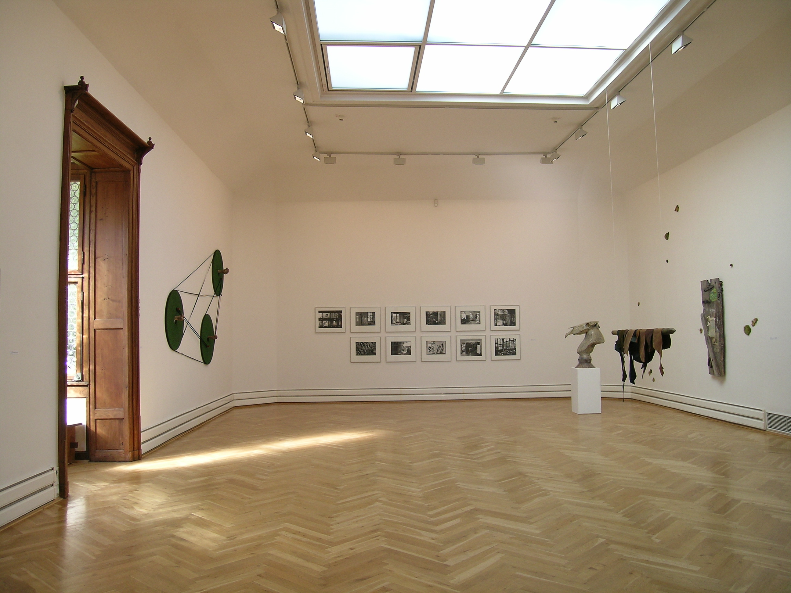 KI generiert: Das Bild zeigt eine helle Kunstgalerie mit mehreren modernen Kunstwerken an den Wänden, einschließlich Fotografien und Installationen. Ein großes Oberlicht und ein Holzboden verstärken die minimalistische Einrichtung des Raums.