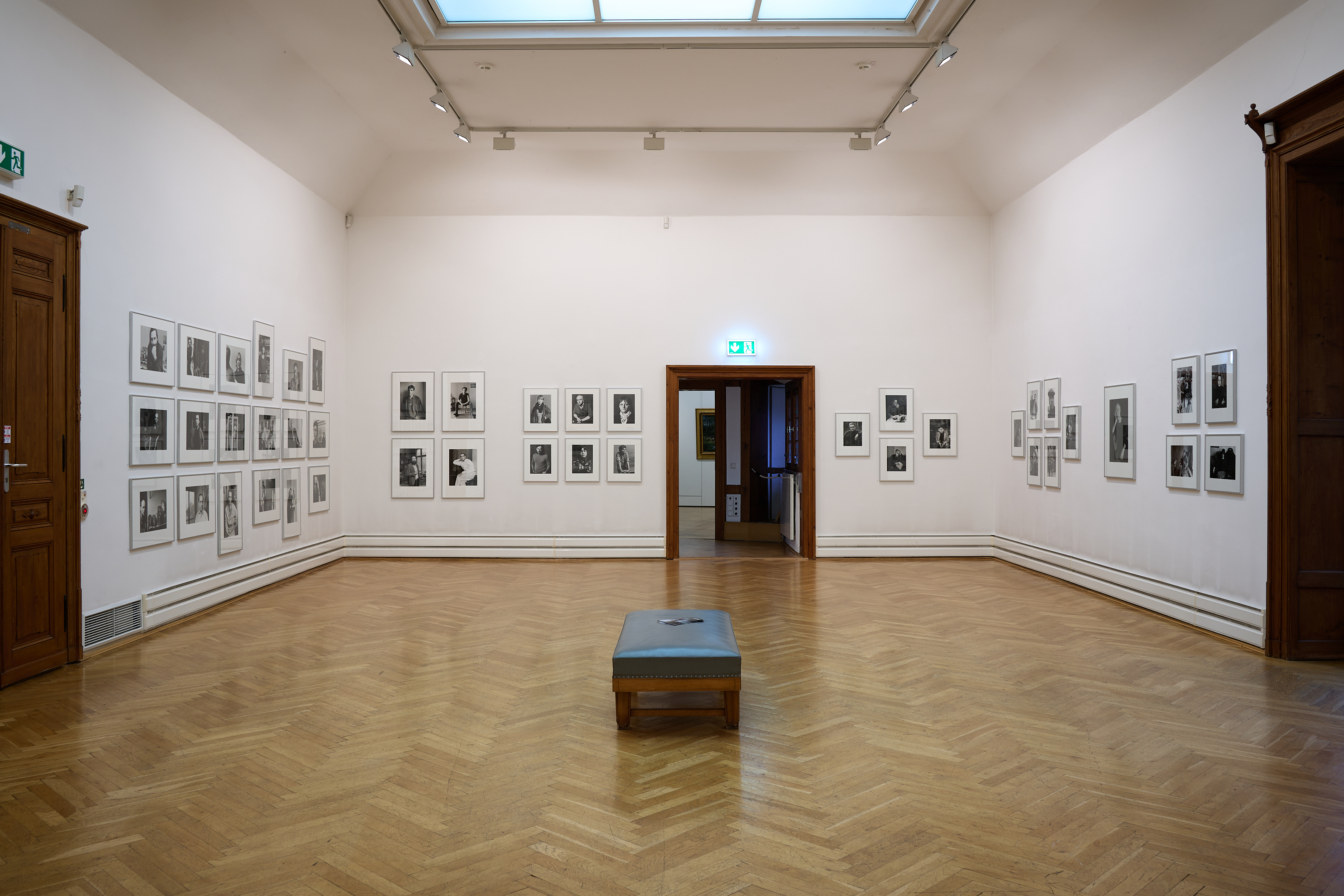 KI generiert: Das Bild zeigt einen Ausstellungsraum in einer Kunstgalerie, in dem zahlreiche Fotografien an den Wänden hängen. In der Mitte des Raumes befindet sich eine Sitzbank und der Raum wirkt ruhig und einladend.