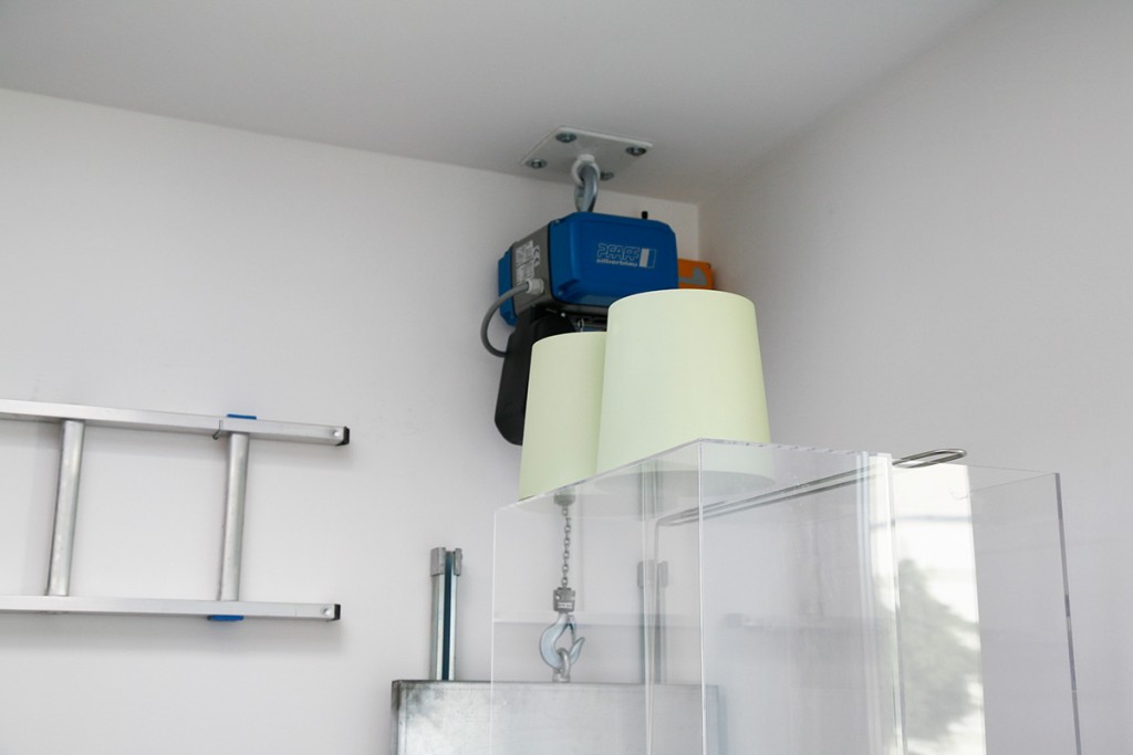 KI generiert: Das Bild zeigt eine Winde oder Seilzug, der an der Decke montiert ist, zusammen mit einer gestützten Leiter an der Wand. Daneben stehen Abdeckungen oder Schutzeinrichtungen aus transparentem Material.