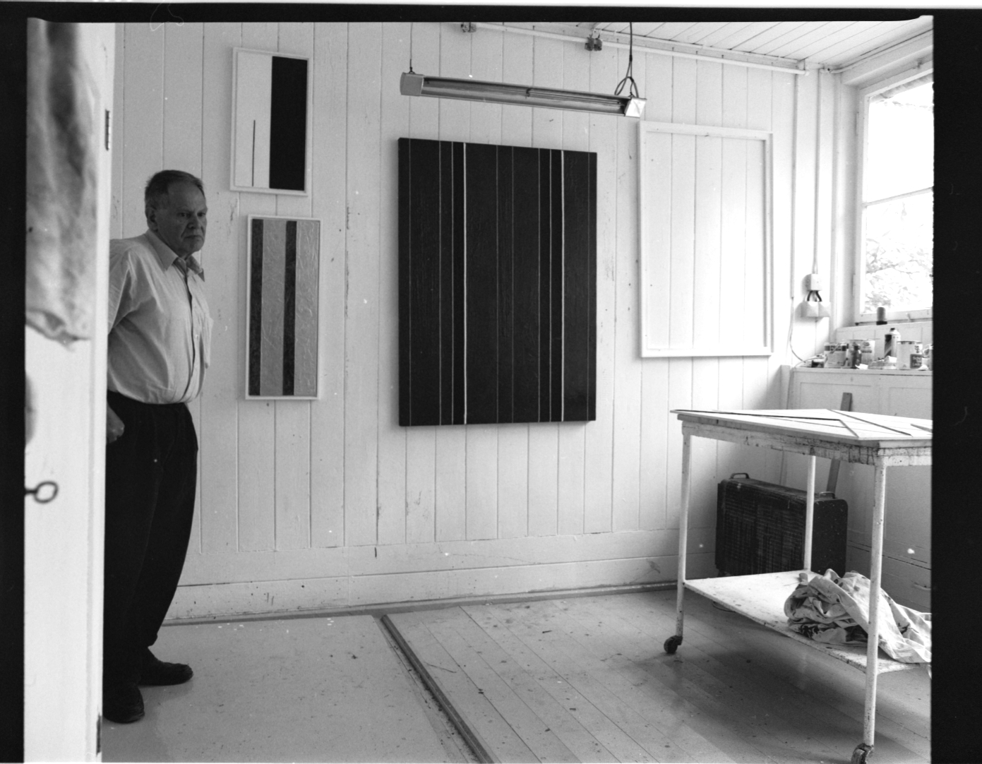 KI generiert: Ein Mann steht in einem minimalistisch eingerichteten Atelier mit mehreren abstrakten Gemälden an der Wand. Der Raum ist hell und schlicht mit einem Tisch und einigen Malutensilien.