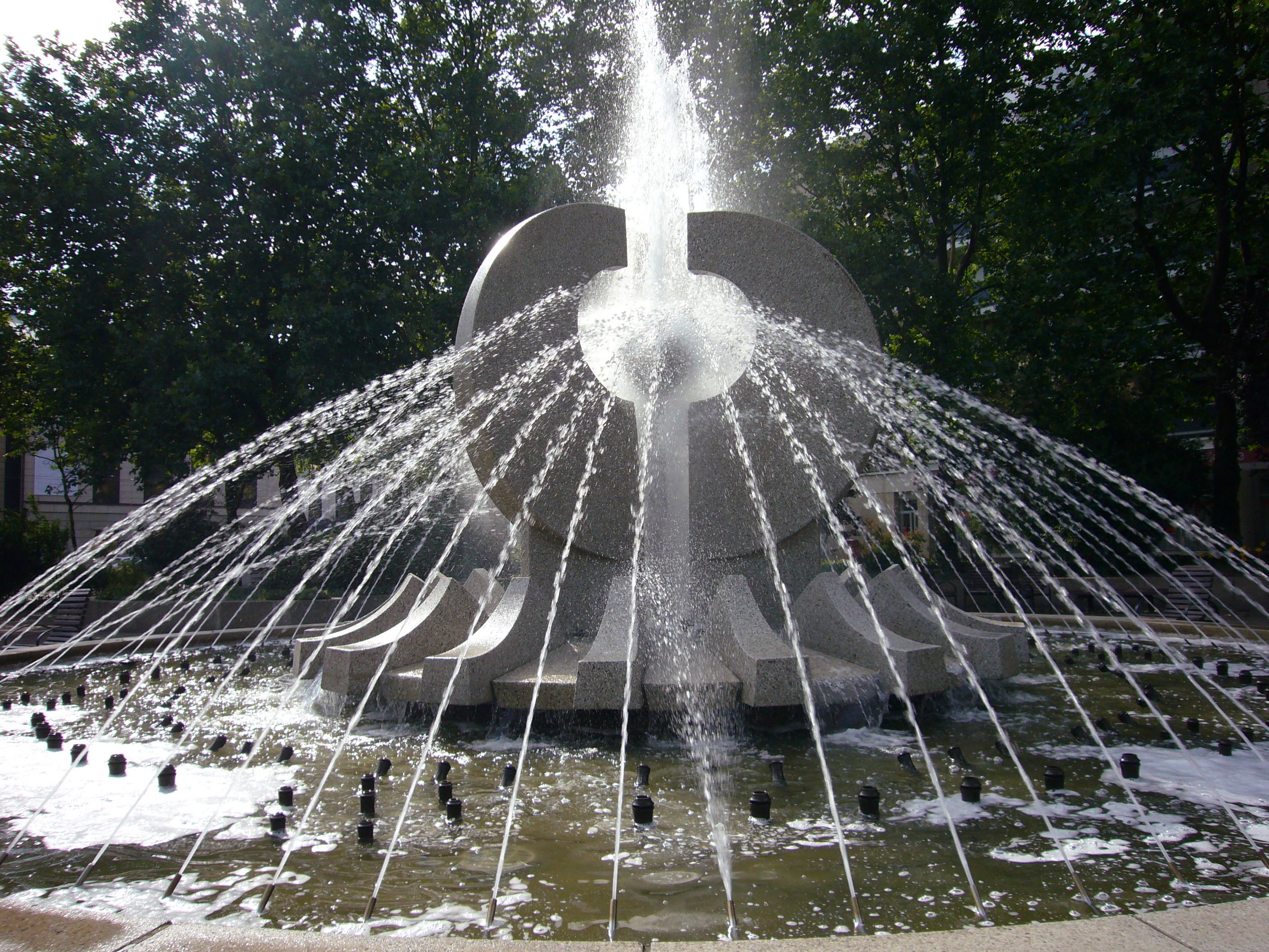 KI generiert: Das Bild zeigt einen Springbrunnen mit einer skulpturalen Struktur, aus der Wasser strömt. Der Brunnen ist von Bäumen umgeben und bildet das zentrale Element der Szene.