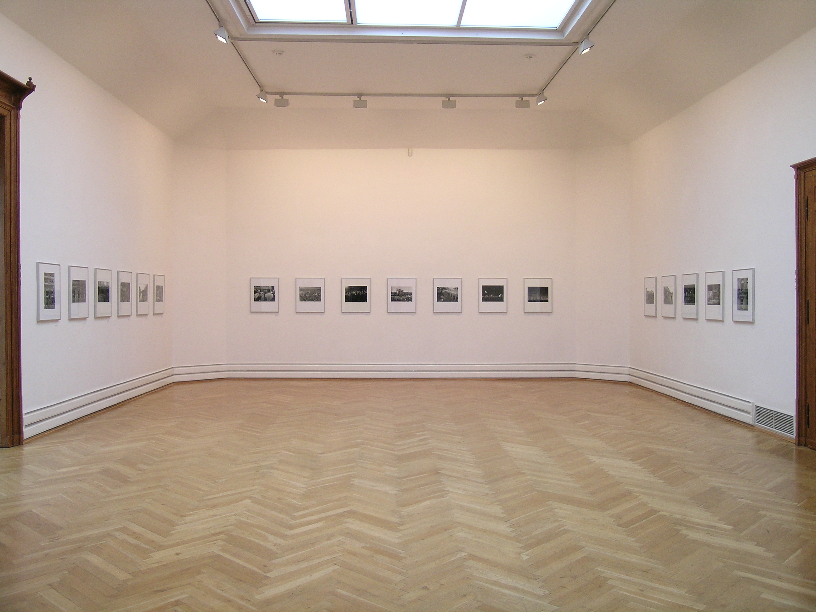 KI generiert: Das Bild zeigt einen weißen Galerieraum mit Holzboden, in dem an den Wänden mehrere gerahmte Fotografien symmetrisch aufgehängt sind. Ein großes Oberlicht beleuchtet den Raum von oben.