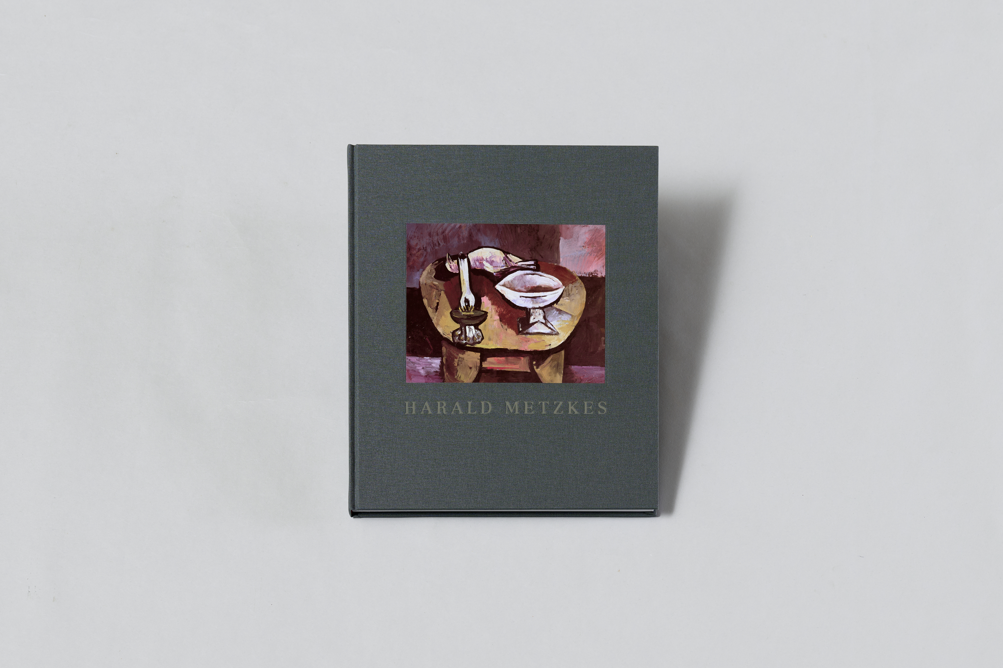 AI generated: The image shows a grey book with the inscription "Harald Metzkes" and a work of art on the cover depicting a table with a candle and a bowl. The artwork is in muted shades of red and brown.