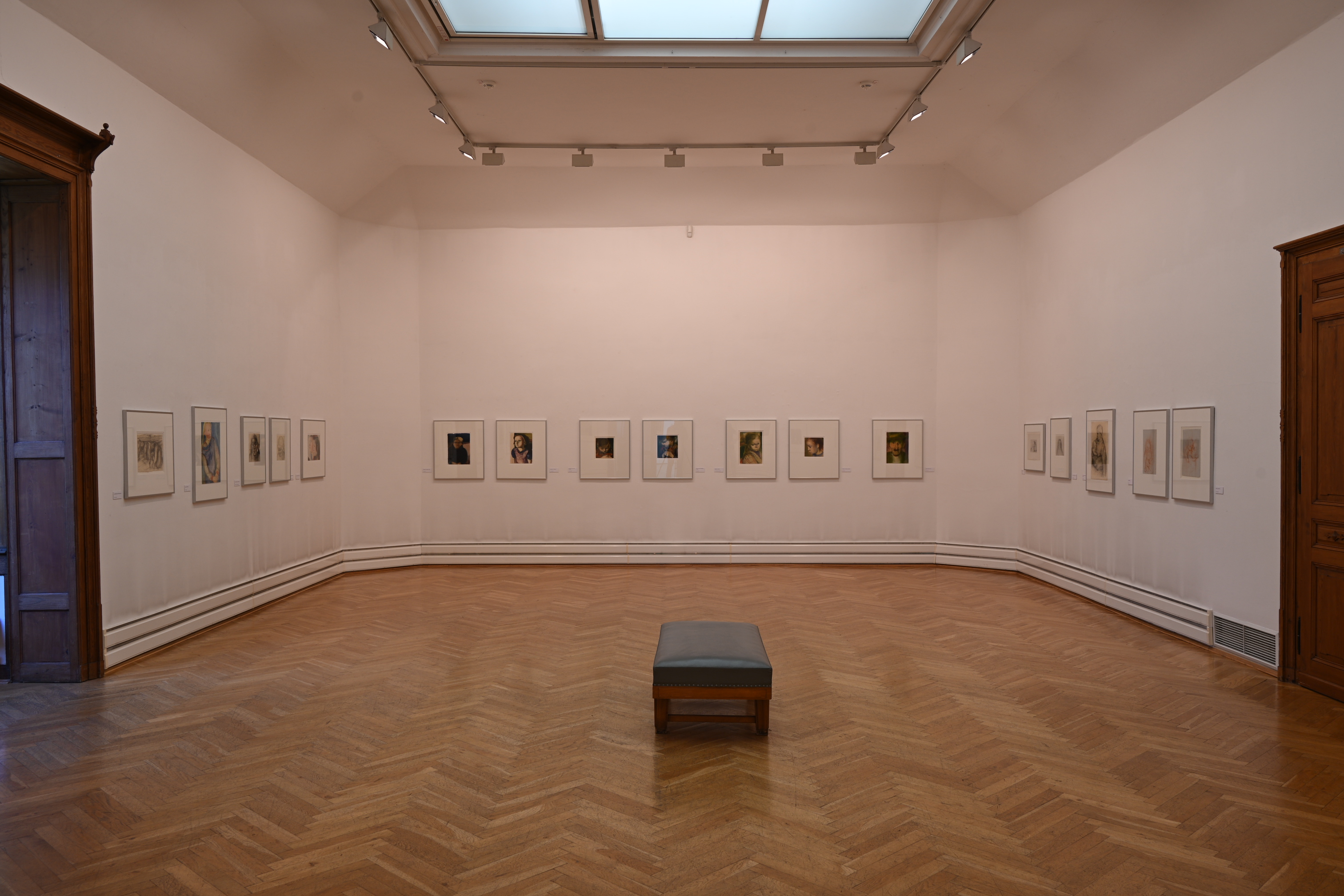 Generated by AI: The image shows an exhibition space in a gallery with white walls, on which numerous framed pictures are hanging. In the centre of the room stands a simple bench on a parquet floor.
