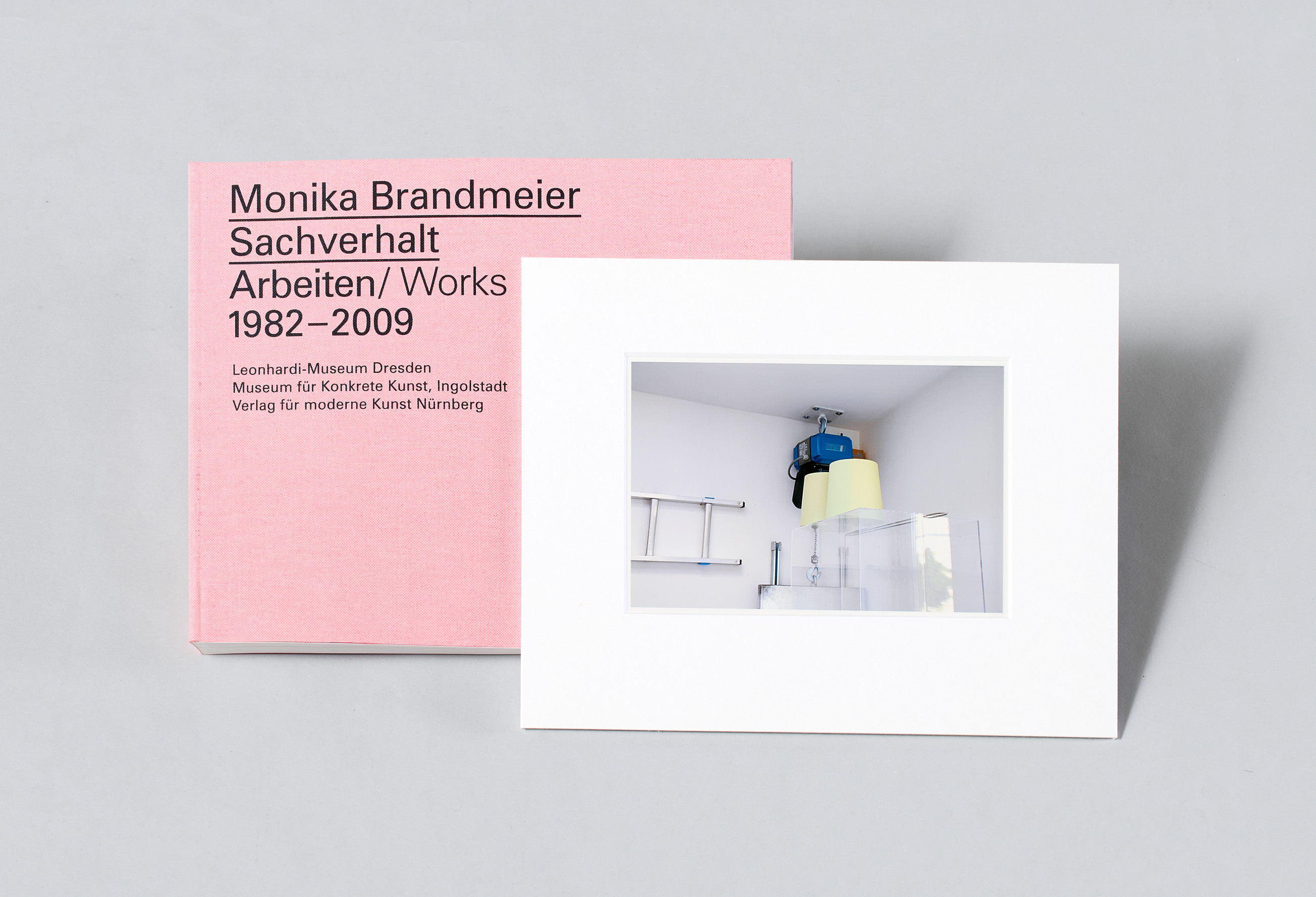 AI generated: The image shows a book with the title "Monika Brandmeier Sachverhalt Arbeiten/Works 1982-2009" in a pink cover, next to it is a white passe-partout with a photo. The photo shows a minimalist work of art or an installation in a room.