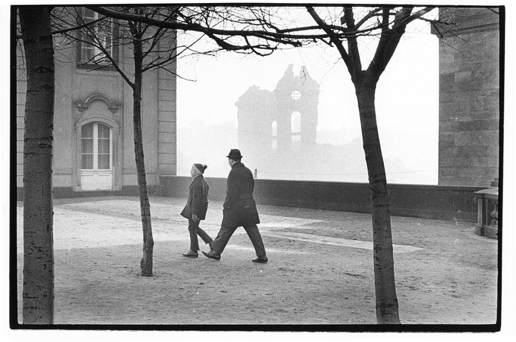 KI generiert: Das Bild zeigt zwei Personen, die in einer städtischen Umgebung spazieren gehen, umgeben von Bäumen. Im Hintergrund sind Gebäuderuinen sichtbar, was eine melancholische oder historische Atmosphäre erzeugt.