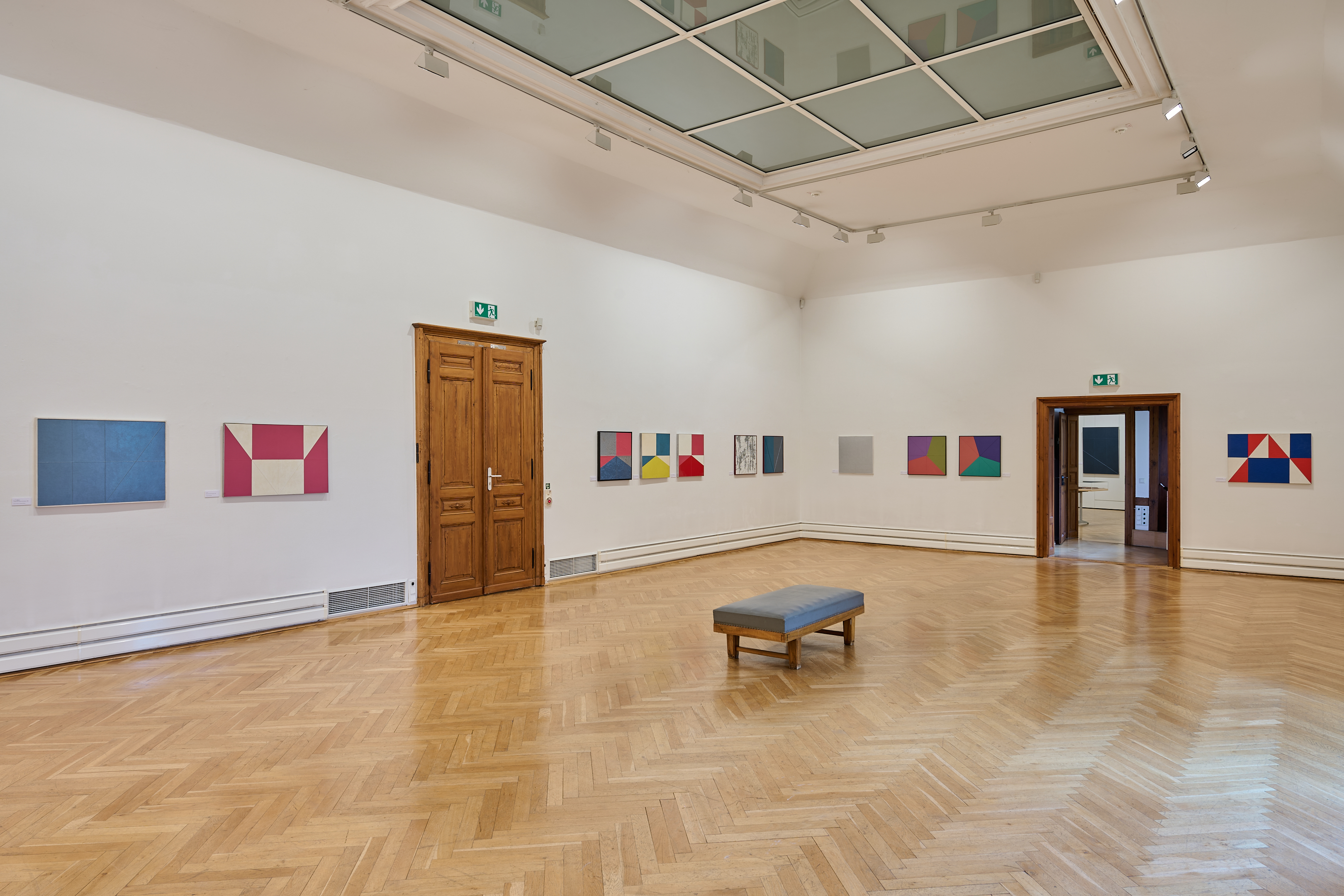 KI generiert: Das Bild zeigt einen modernen Ausstellungsraum in einem Museum oder einer Galerie mit abstrakten Kunstwerken an den Wänden. Der Raum ist geräumig und hat einen hölzernen Boden mit einer einfachen Bank in der Mitte.