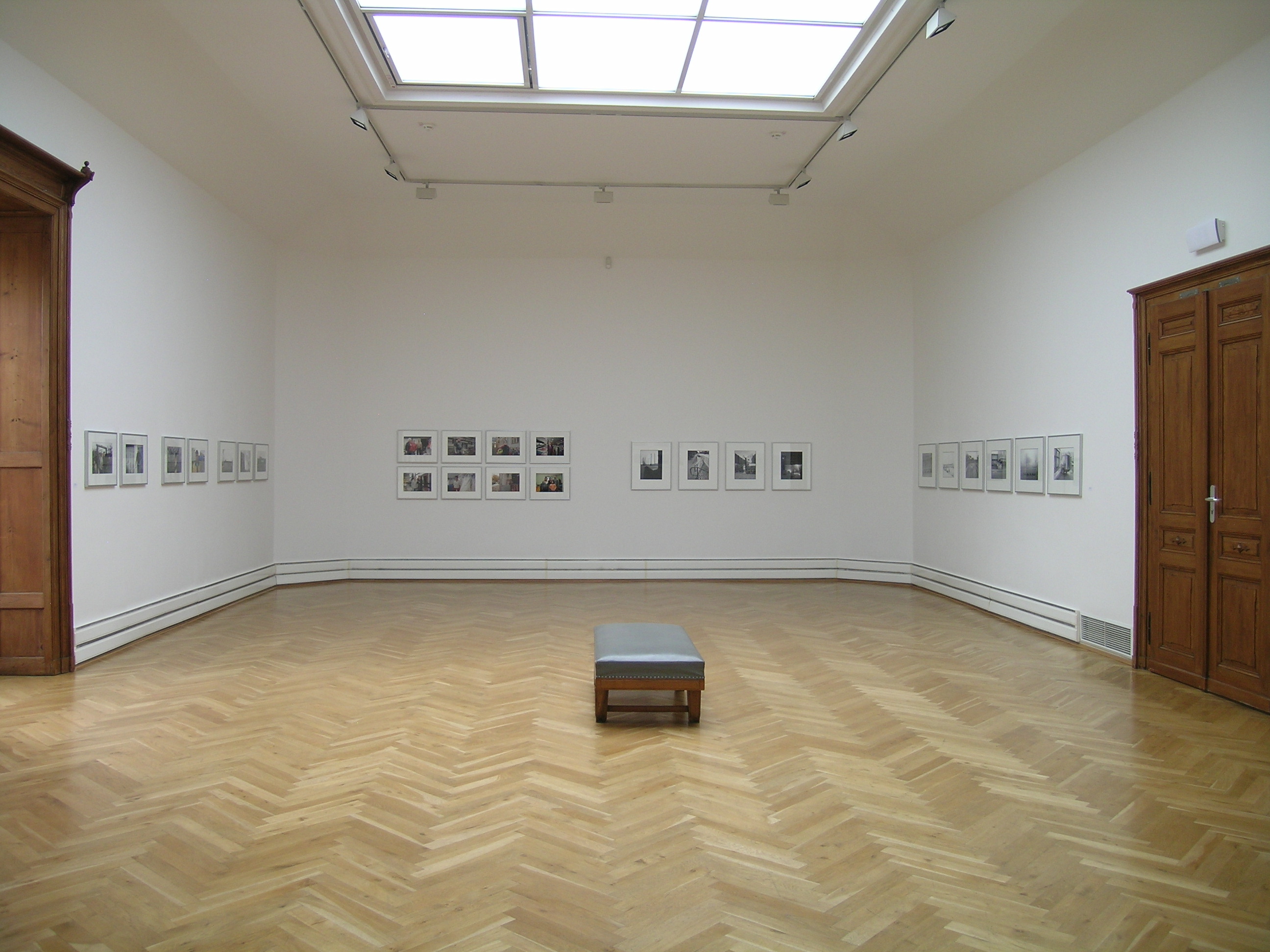 KI generiert: Das Bild zeigt einen leeren Museumsraum mit Holzboden und weiß gestrichenen Wänden, an denen gerahmte Kunstwerke hängen. Im Zentrum des Raumes befindet sich eine graue Sitzbank.