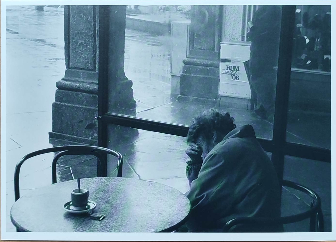 KI generiert: Das Bild zeigt eine Person, die in einem Café an einem Tisch sitzt und den Kopf in die Hand stützt. Auf dem Tisch befindet sich eine Tasse, und draußen scheint es regnerisch zu sein.